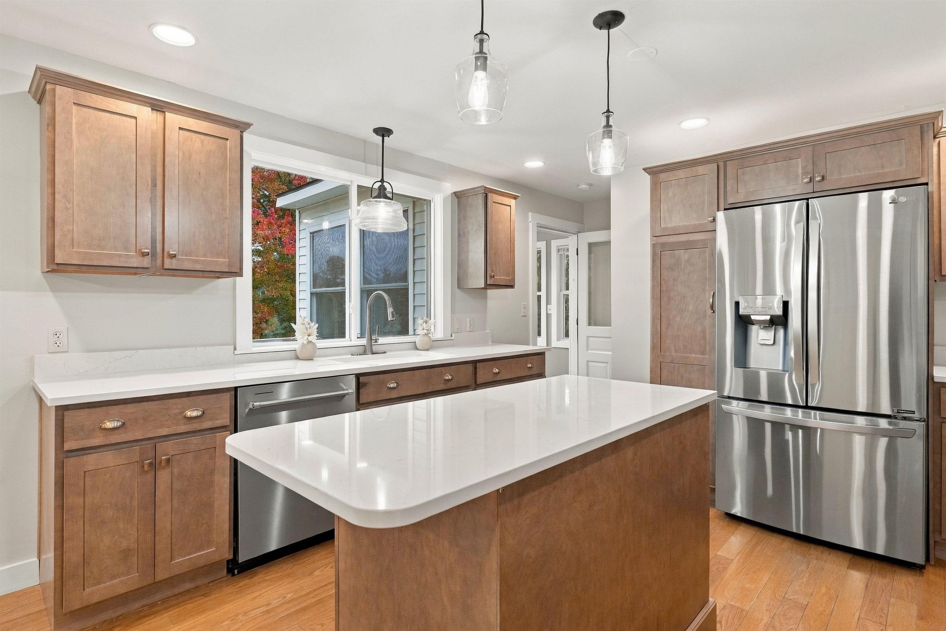 Kitchen with a large white-counter island, warm wood cabinets, stainless steel appliances, and autumn foliage outside.