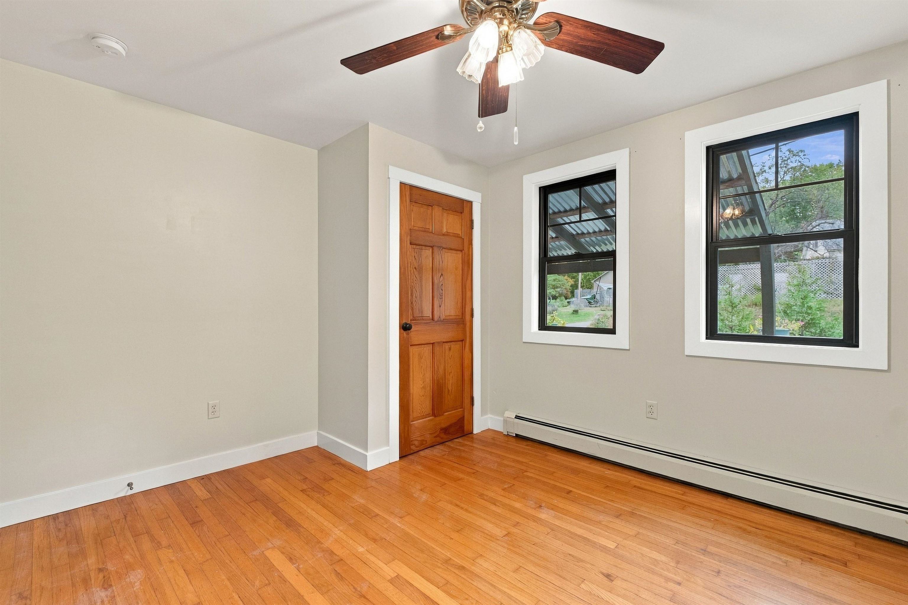 Empty room with hardwood floors, a wooden door, two windows, and a ceiling fan.