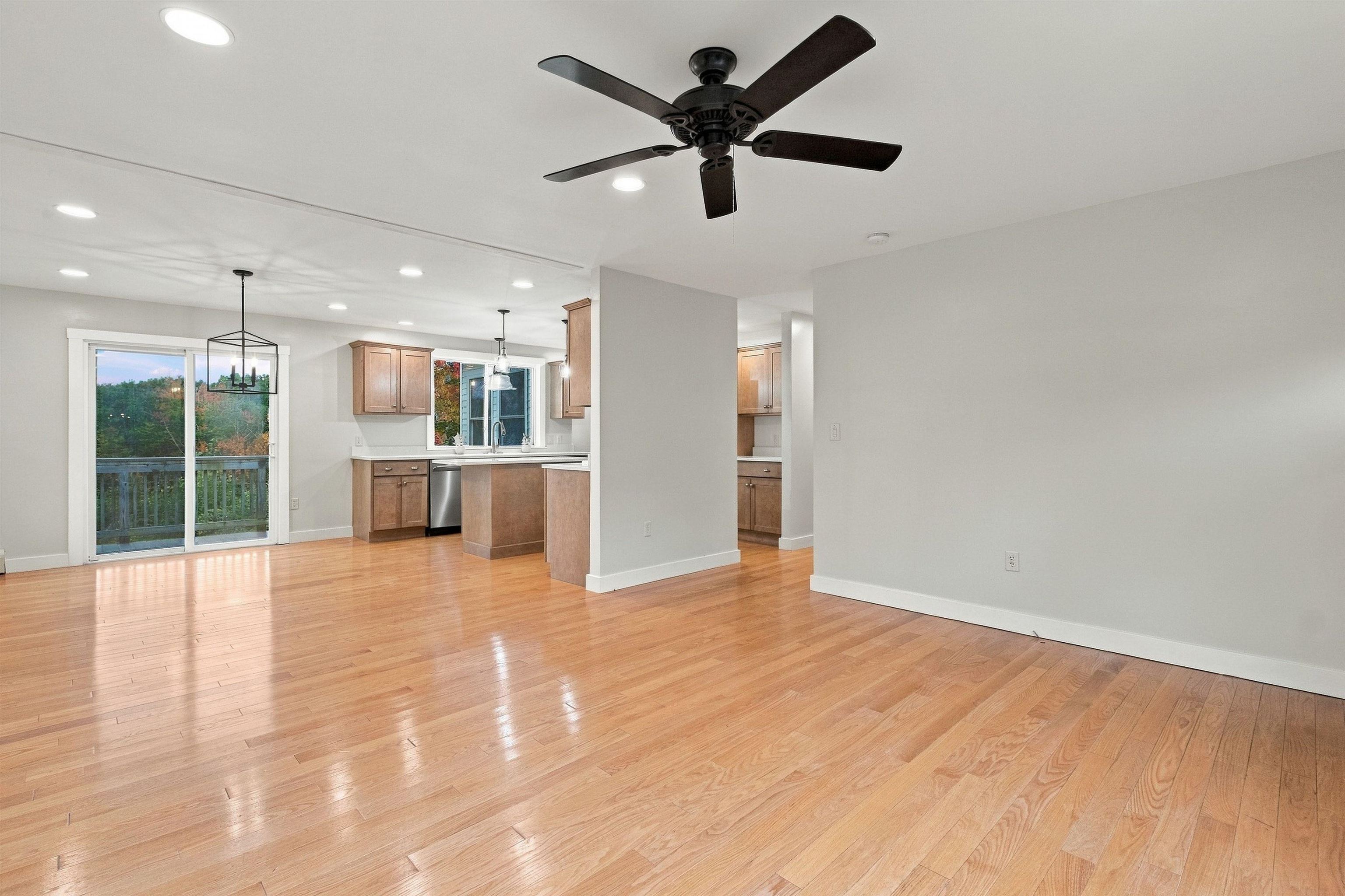 Empty open-plan living and dining area with hardwood floors, kitchen, ceiling fan, and sliding doors to a wooded deck.
