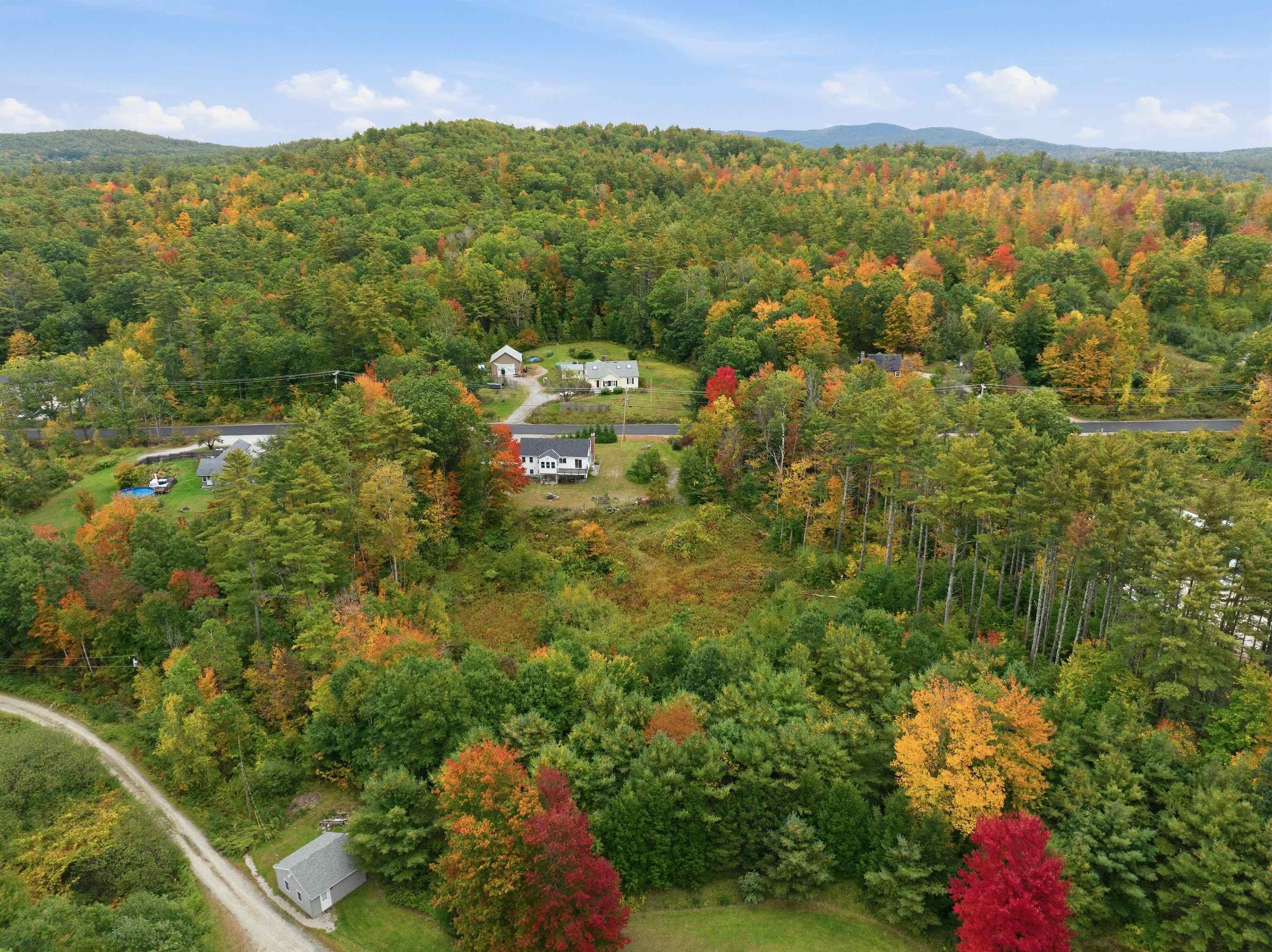 Aerial view of a home amid autumn forest, with a road and distant wooded hills.