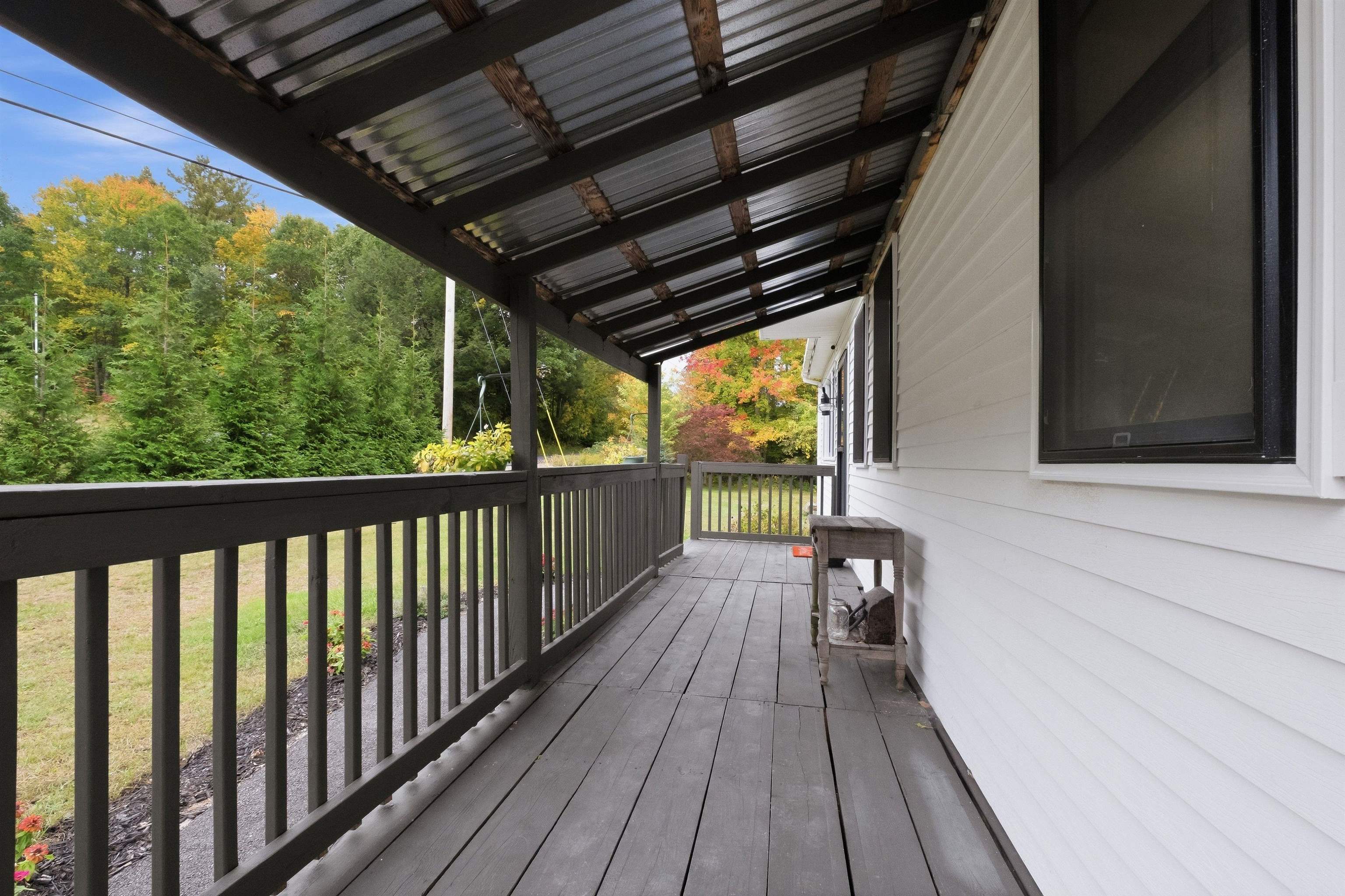 Covered porch with dark railings and decking beside white siding, overlooking colorful autumn trees.