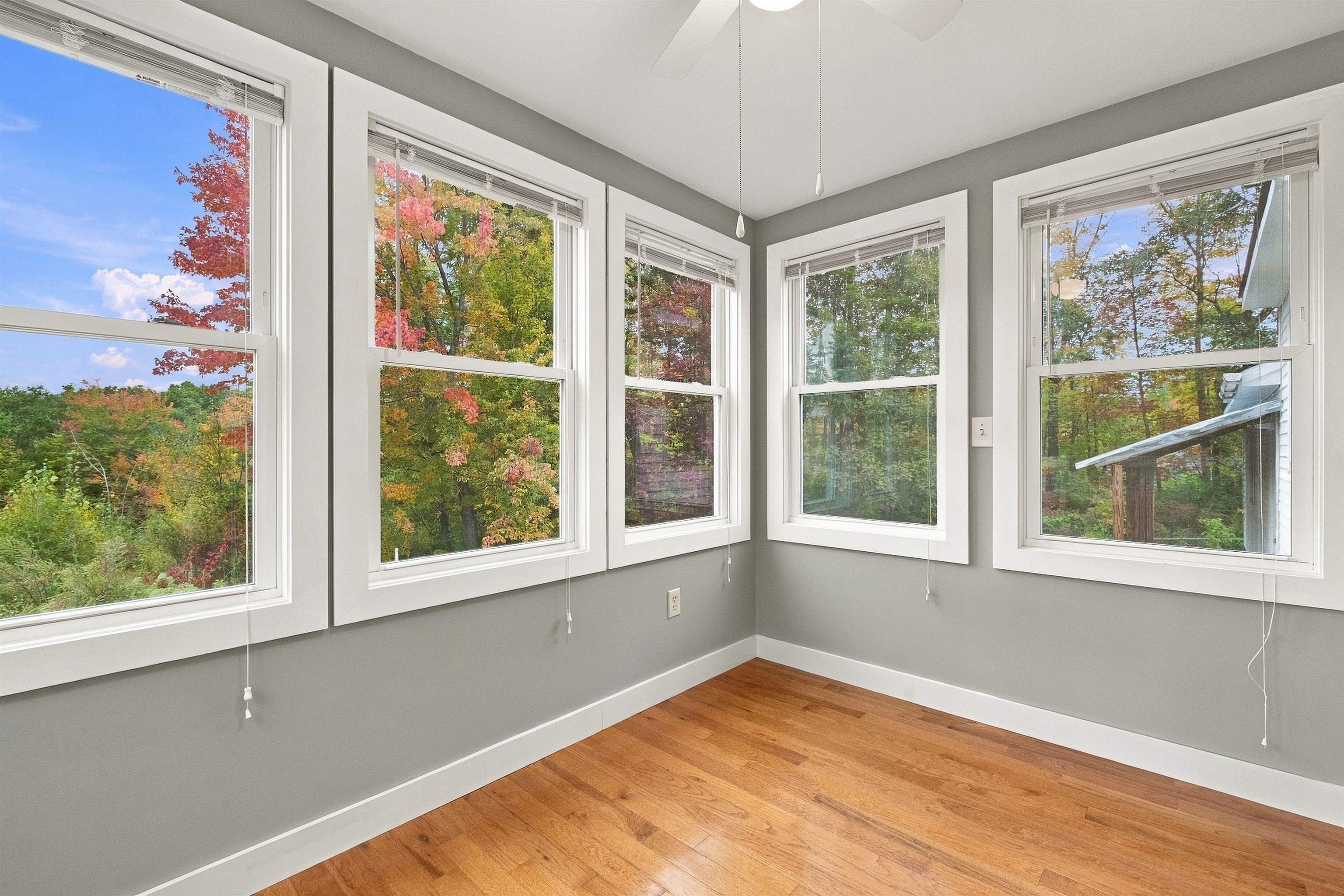 Vacant room with hardwood floors and large windows overlooking autumn foliage.