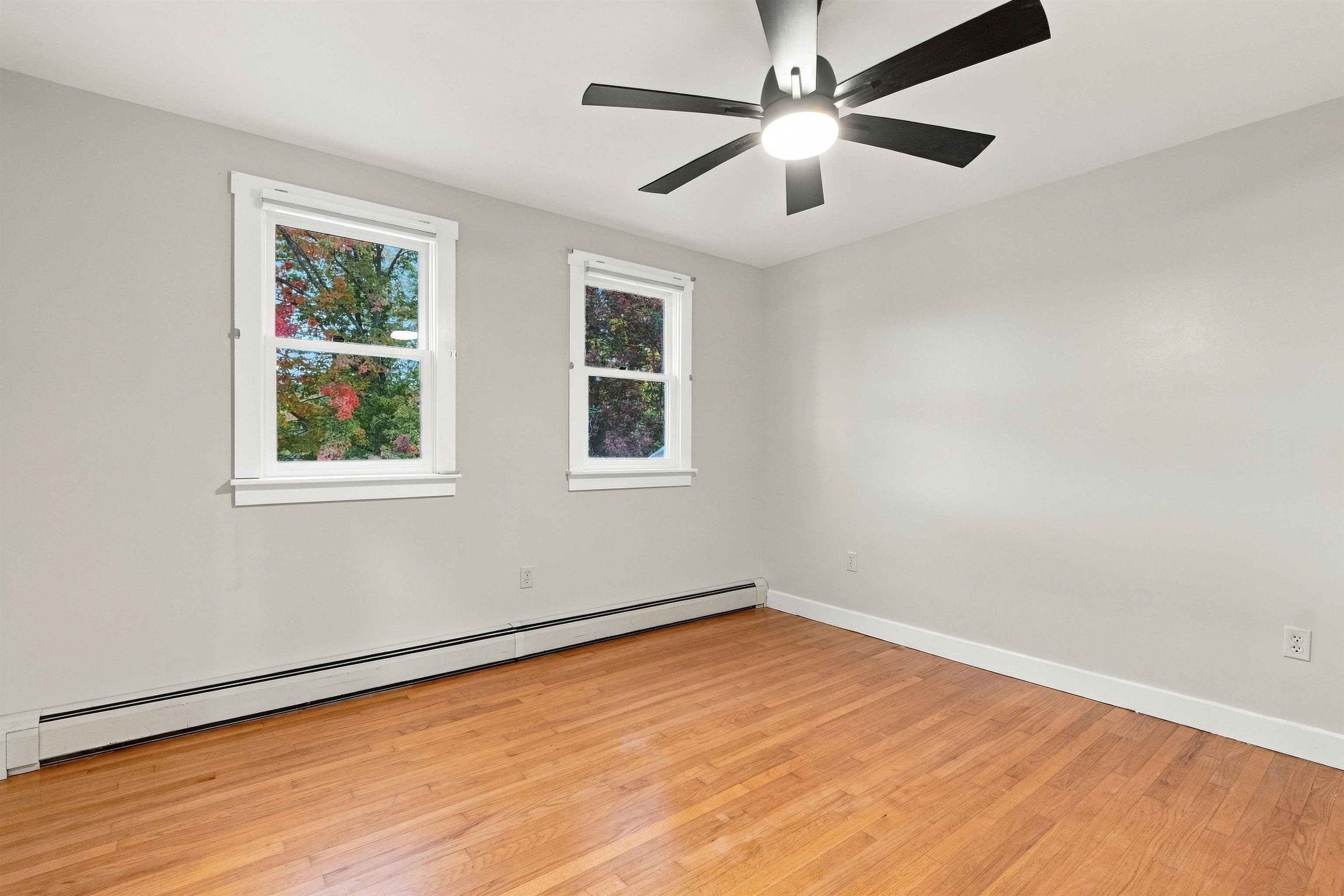 Empty room with hardwood floors, two windows, a ceiling fan, and a long baseboard heater.