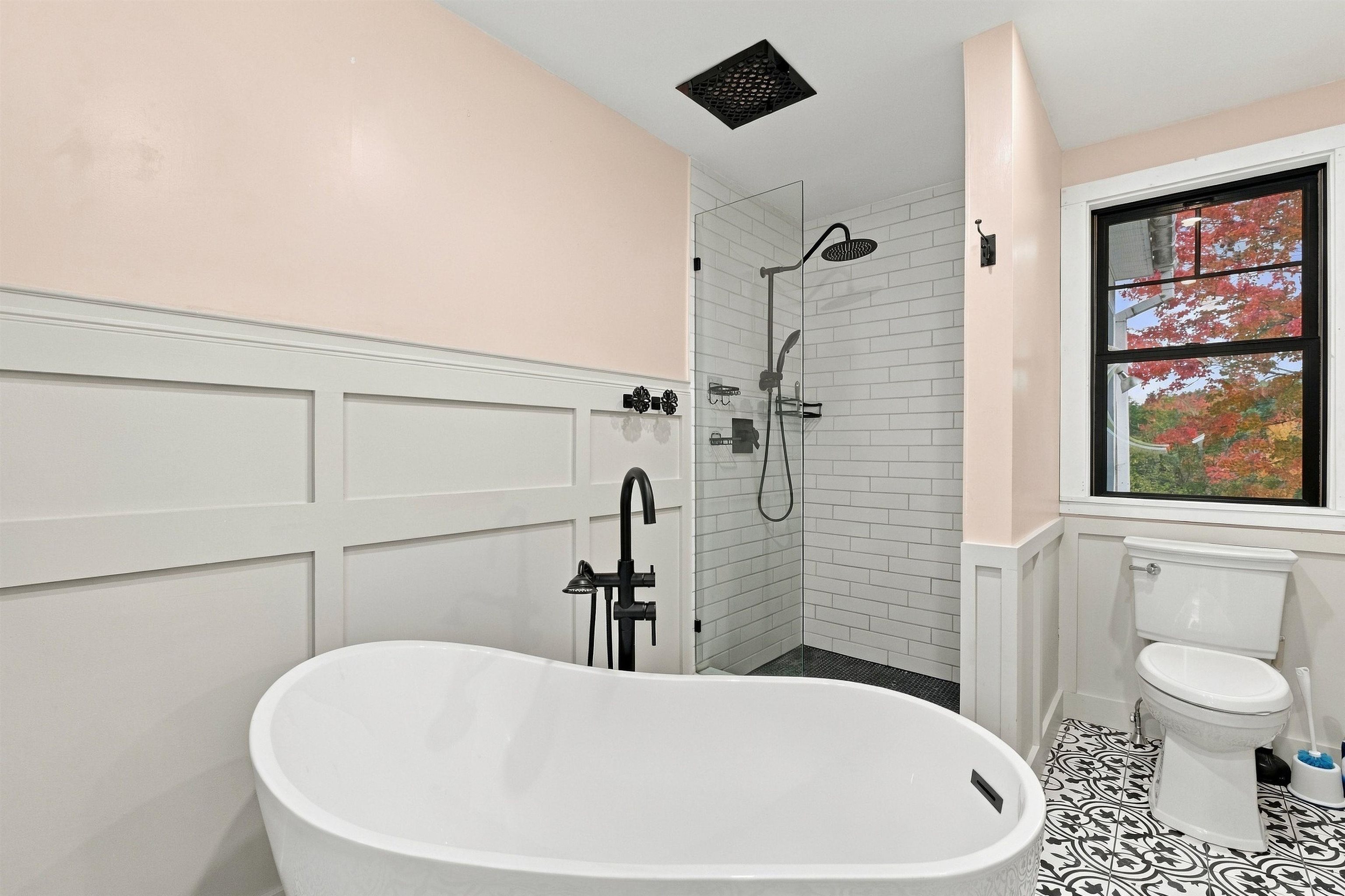 Bathroom with a freestanding tub, glass shower, patterned floor, and autumn foliage visible through the window.