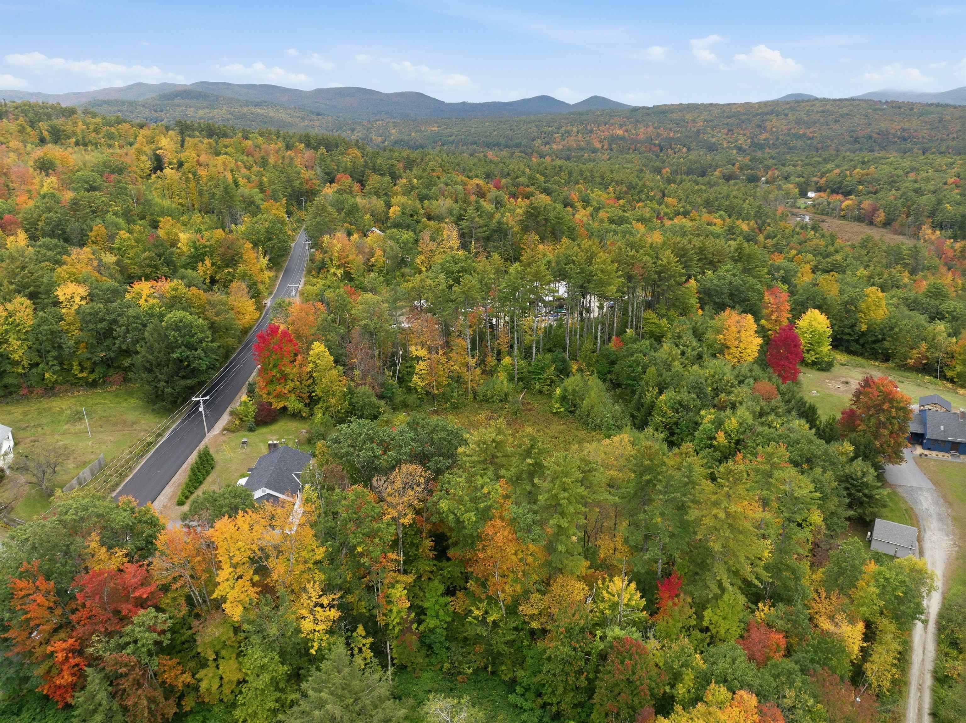 Aerial view of autumn woods, a winding road, scattered homes, and distant mountains.