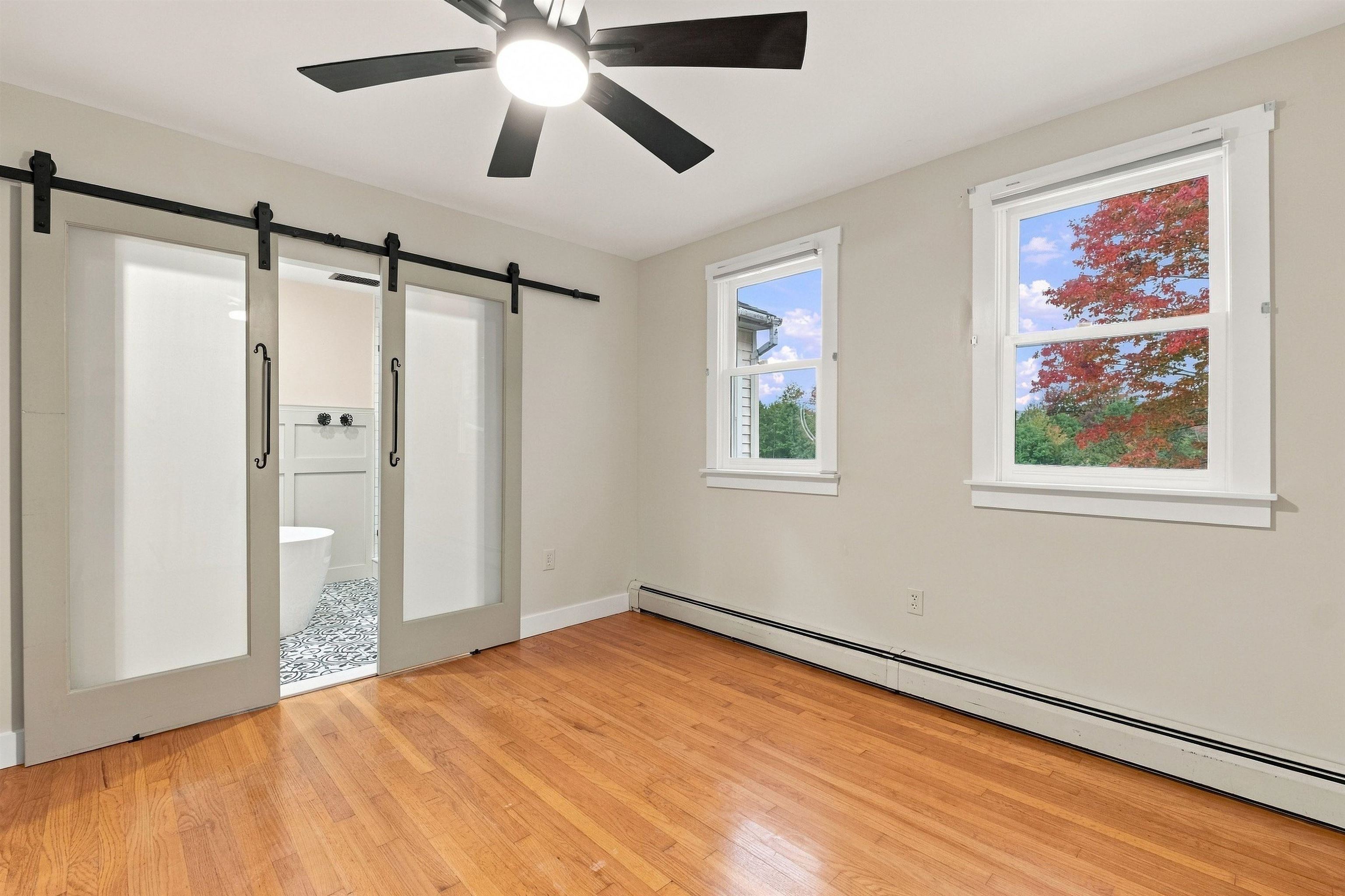 Empty bedroom with hardwood floors, sliding barn doors, and windows framing autumn foliage.