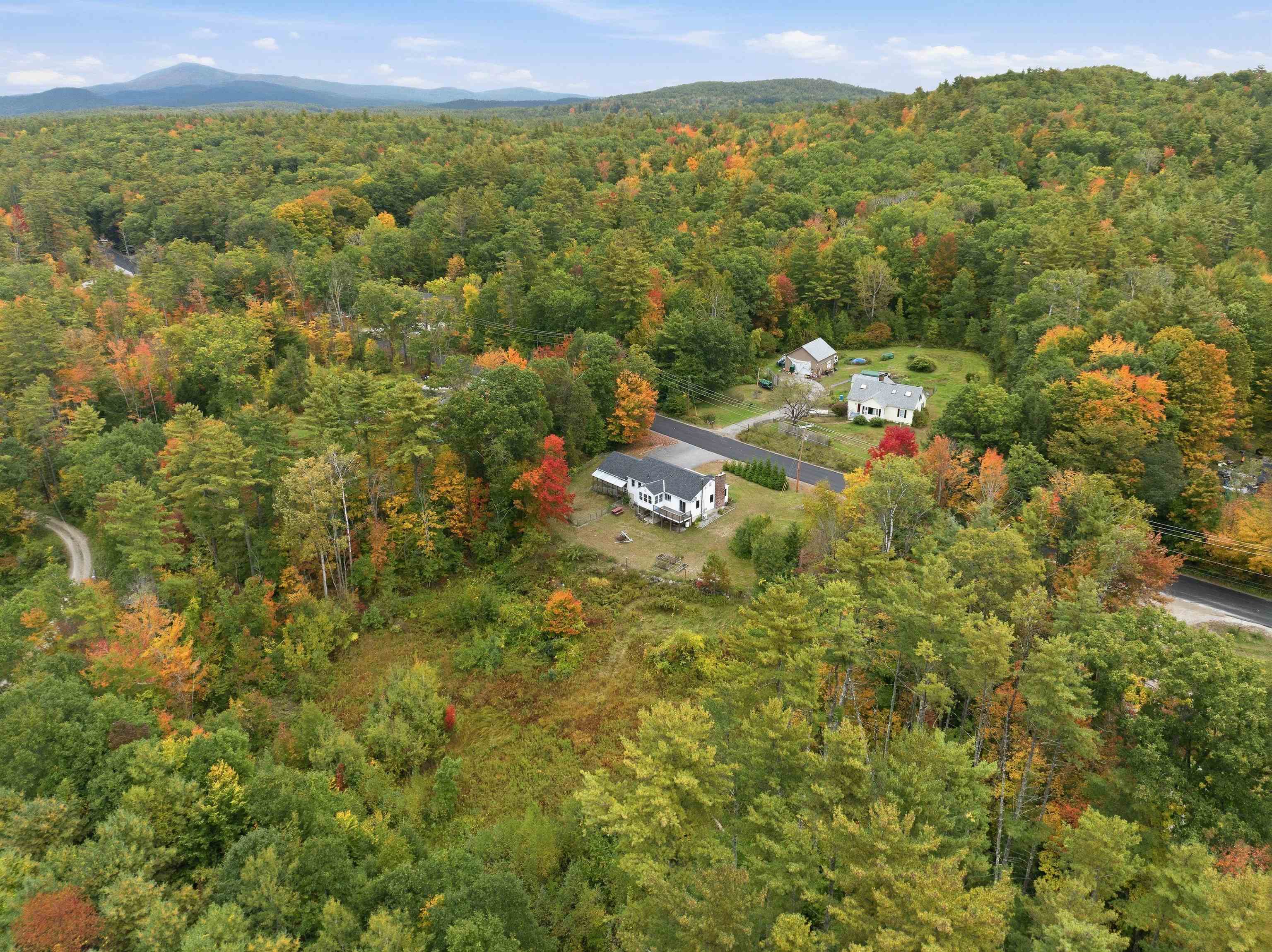 Aerial view of a white house amid autumn woodland, with a nearby road and distant mountains.
