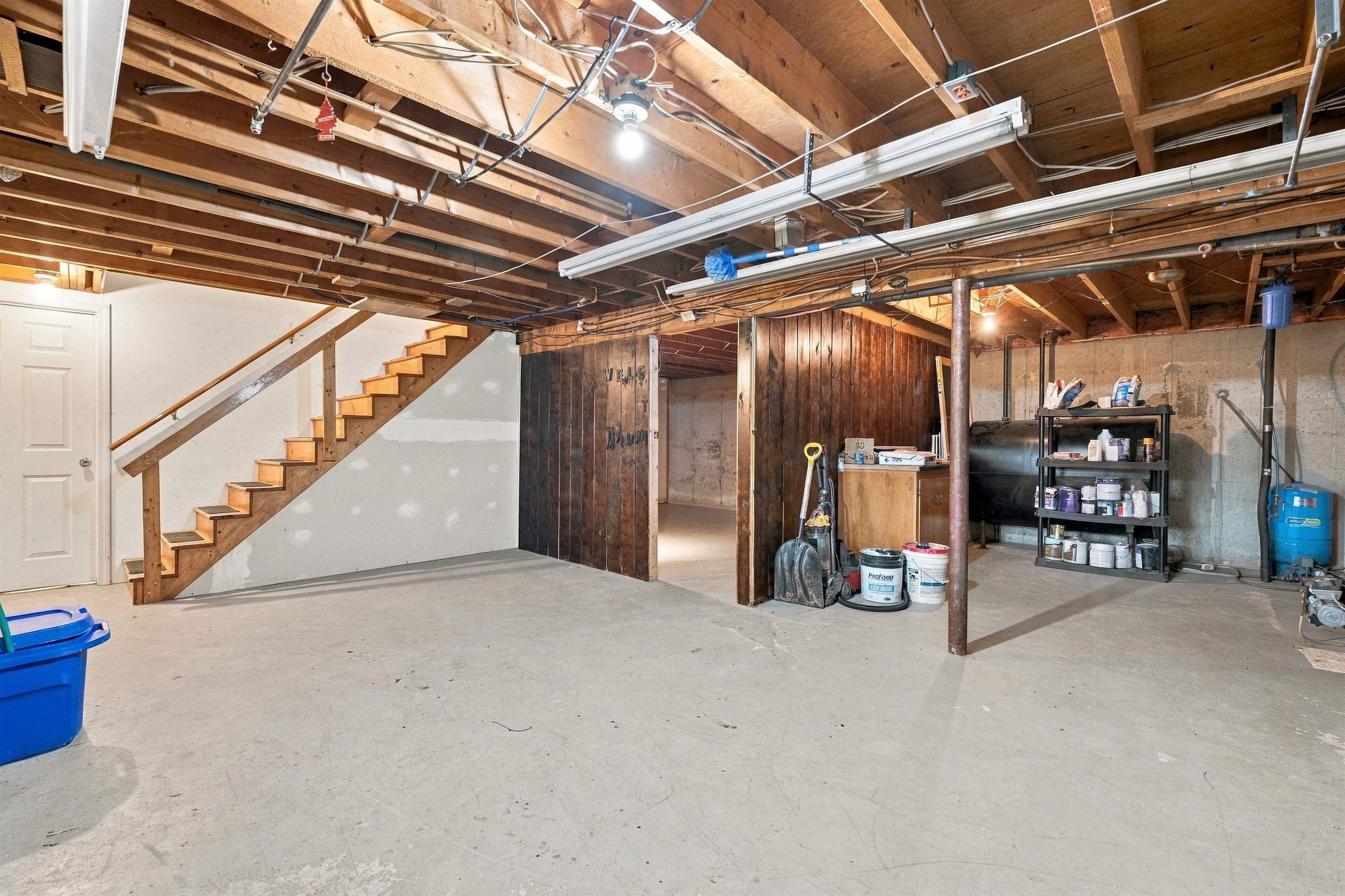 Unfinished basement with exposed ceiling joists, concrete floor, wooden stairs, and storage shelves.