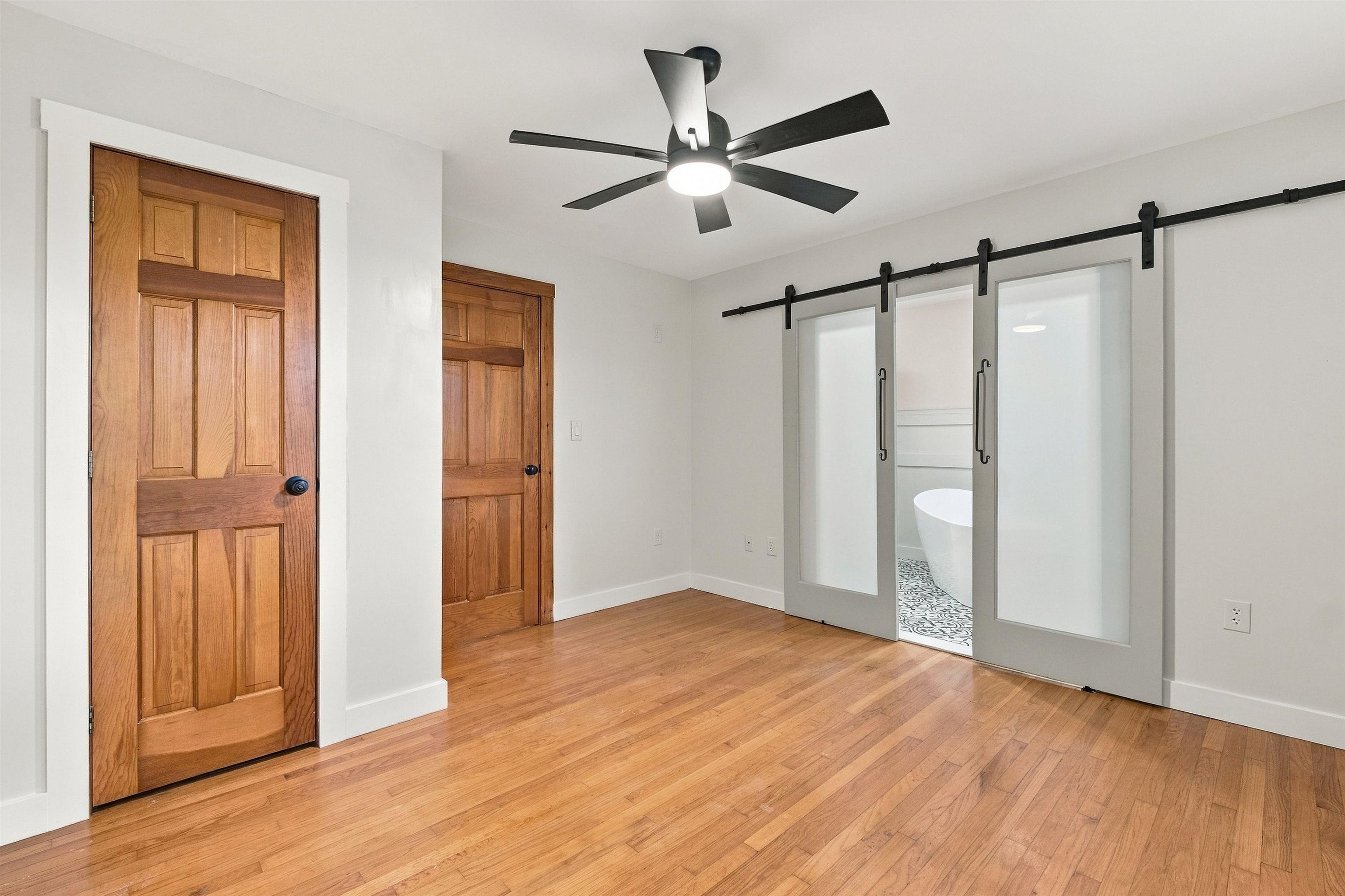 Empty room with hardwood floors, wood doors, and frosted-glass barn doors opening to a bathroom with a freestanding tub.