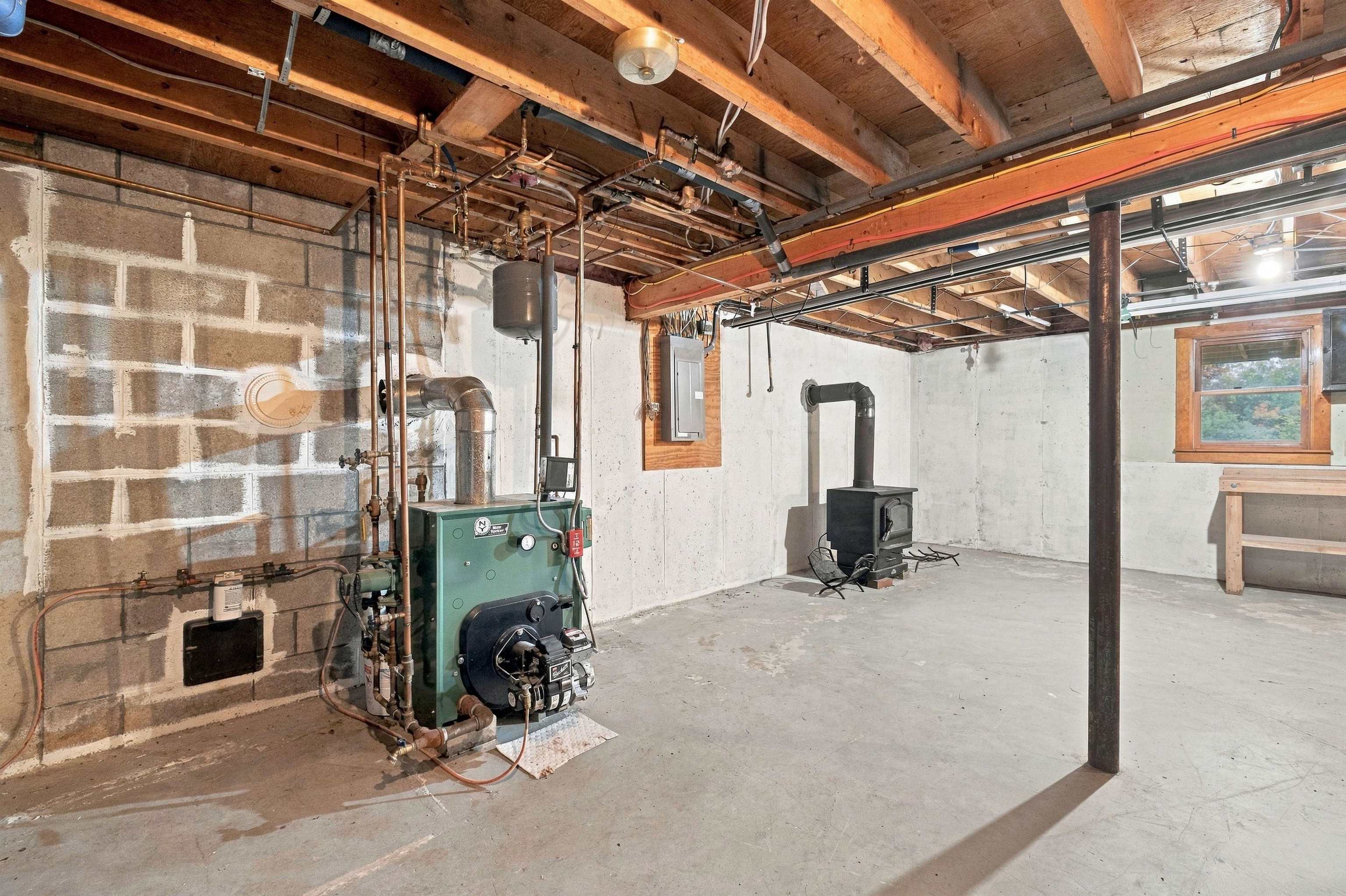 Unfinished basement with a green boiler, exposed pipes, and a black wood stove.
