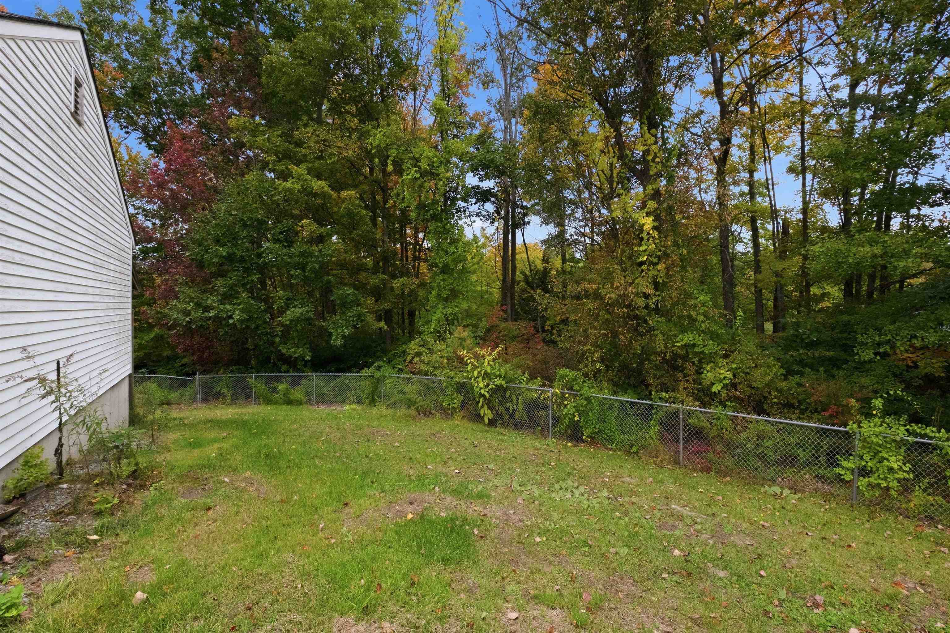 Grassy backyard beside a white-sided house, with a chain-link fence and wooded autumn backdrop.
