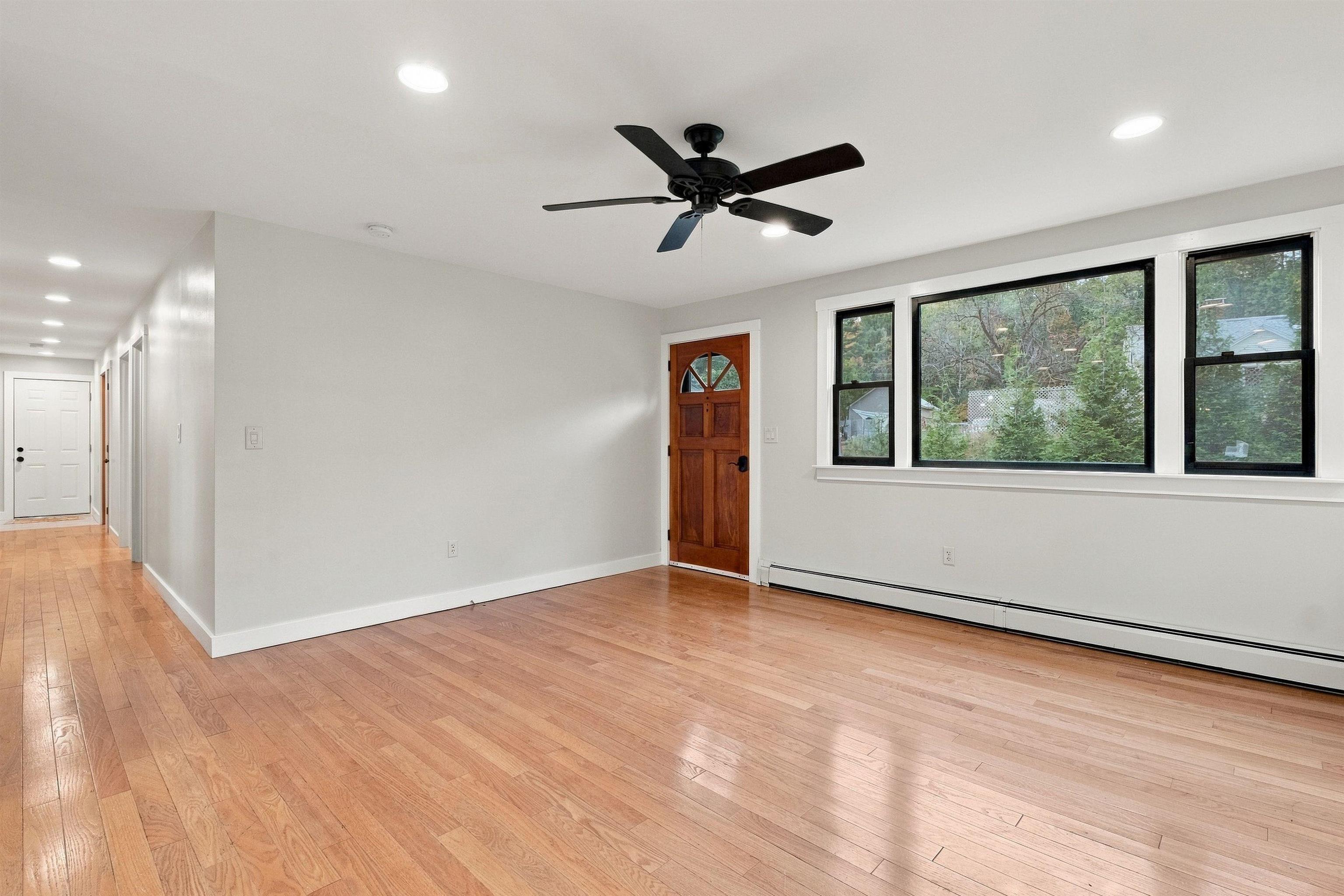 Empty living room with hardwood floors, a ceiling fan, large windows, and a distinctive wood-paneled door.