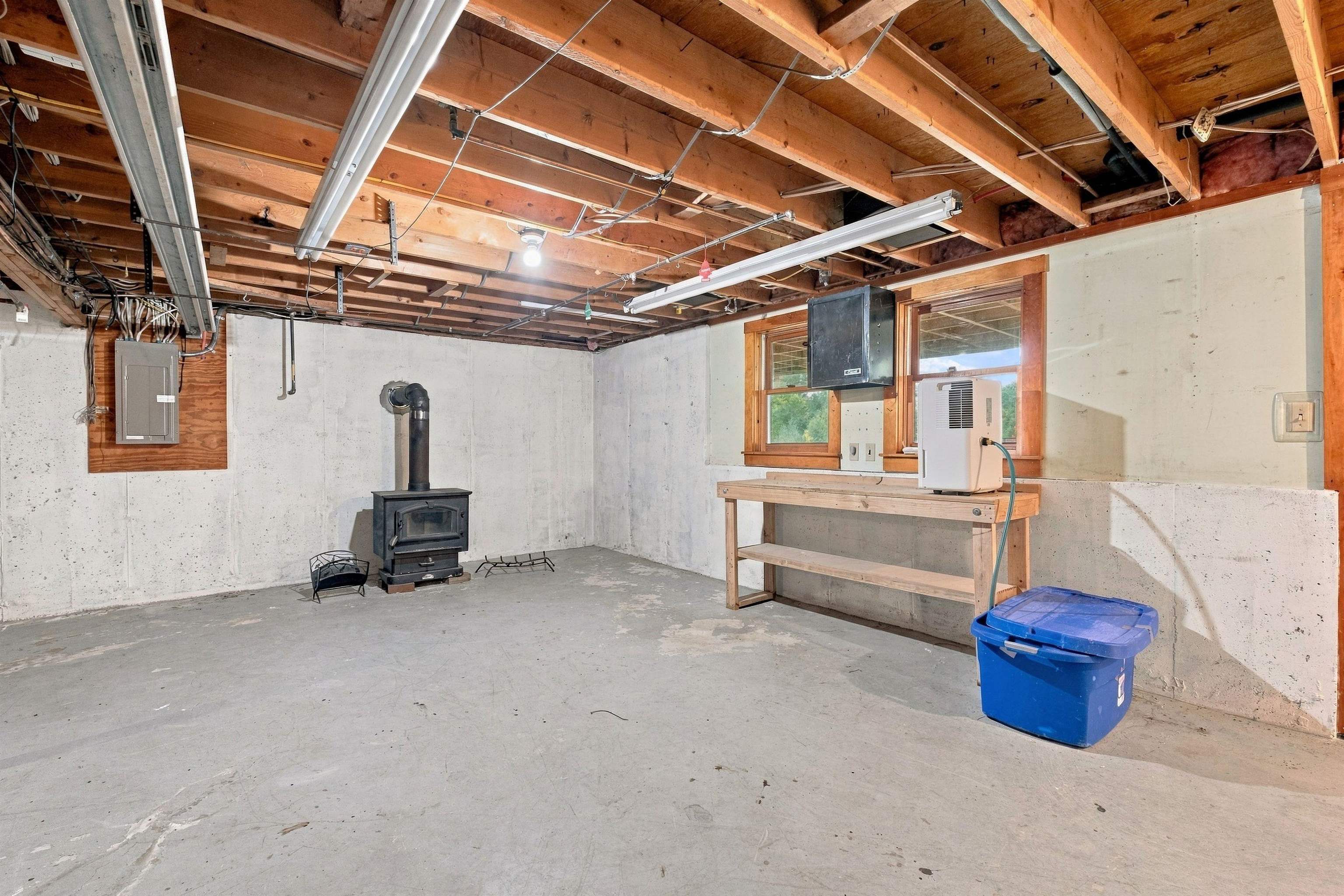Unfinished basement with exposed joists, a wood stove, and a workbench by two windows.