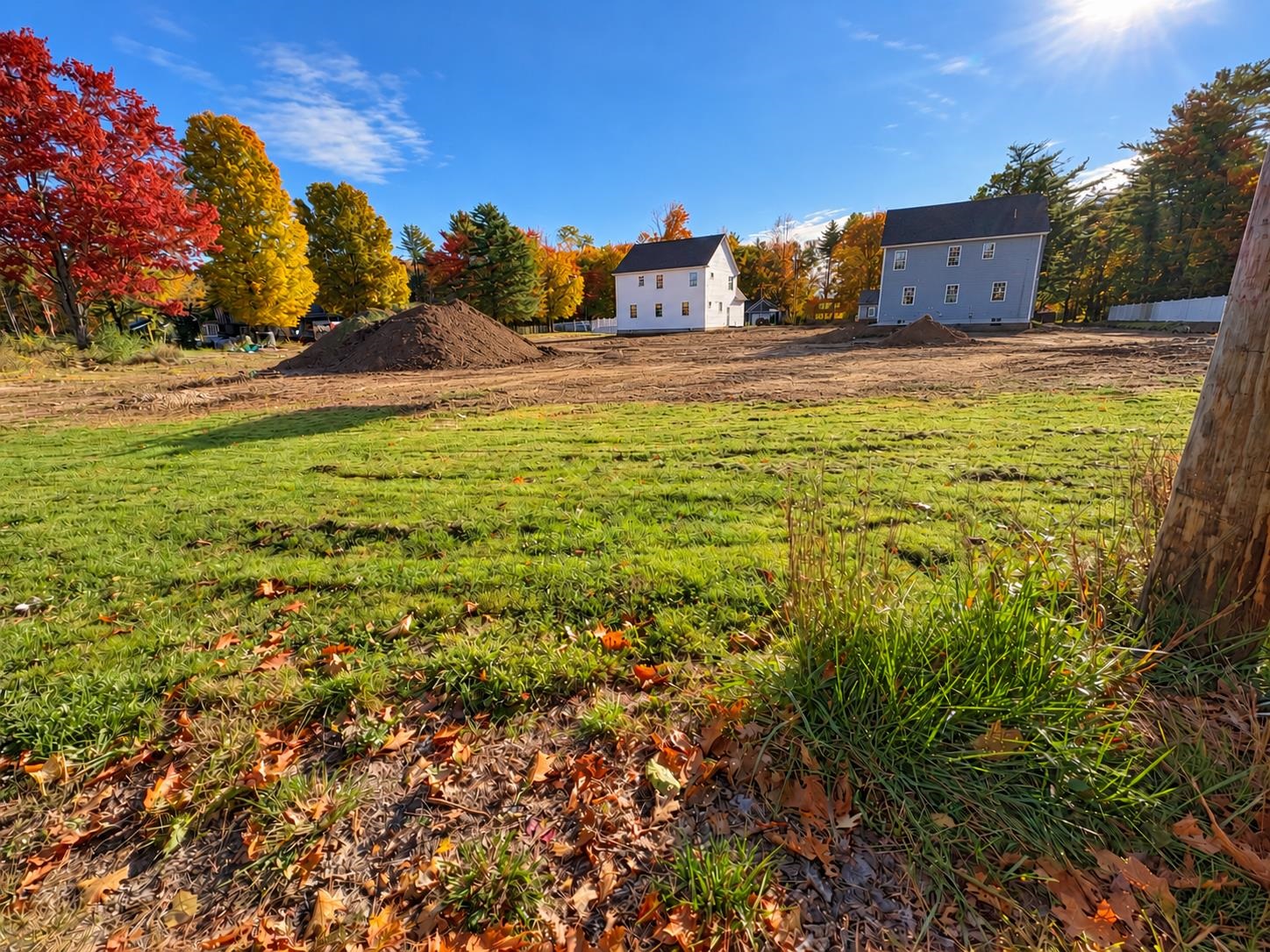 Cleared residential lot with soil piles and two houses amid autumn foliage.