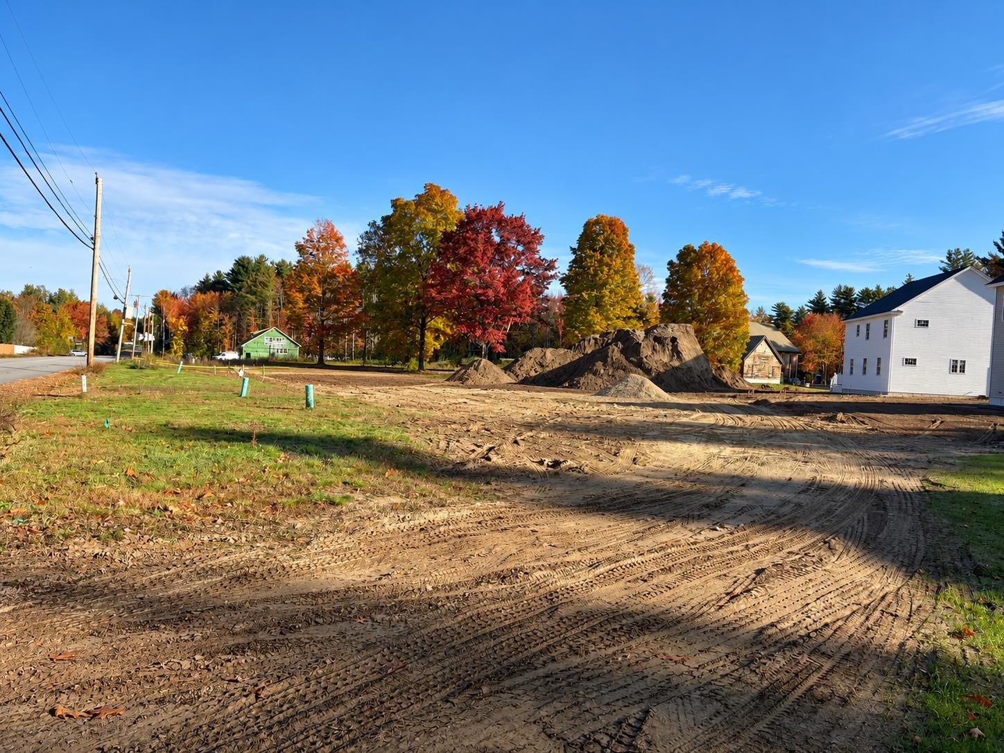 Cleared construction site with graded earth and soil piles beside autumn trees and nearby homes.