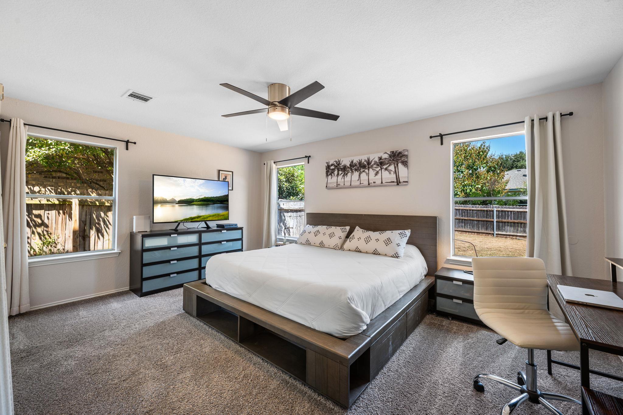 Bedroom with a dark platform bed, white bedding, ceiling fan, and windows overlooking a fenced yard.