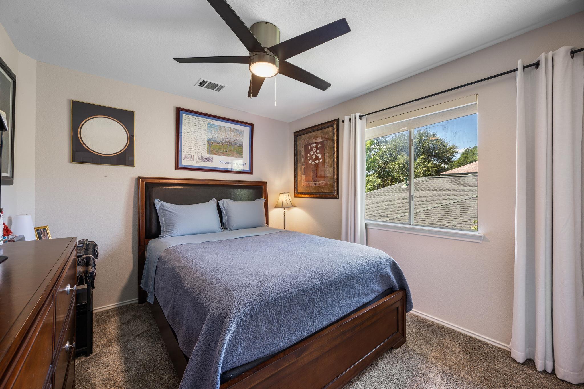 Bedroom with a dark wood bed, blue patterned bedding, ceiling fan, and curtained window.