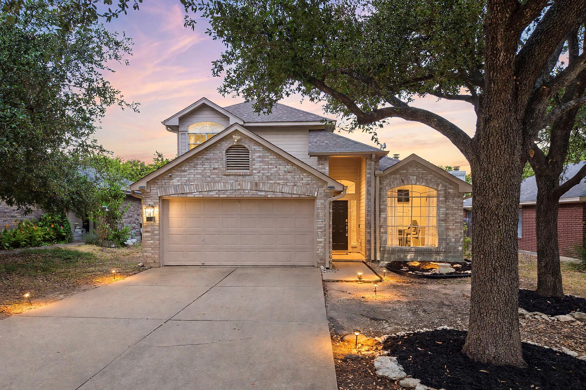 Two-story brick home with a double garage and warmly lit windows beneath mature trees at dusk.