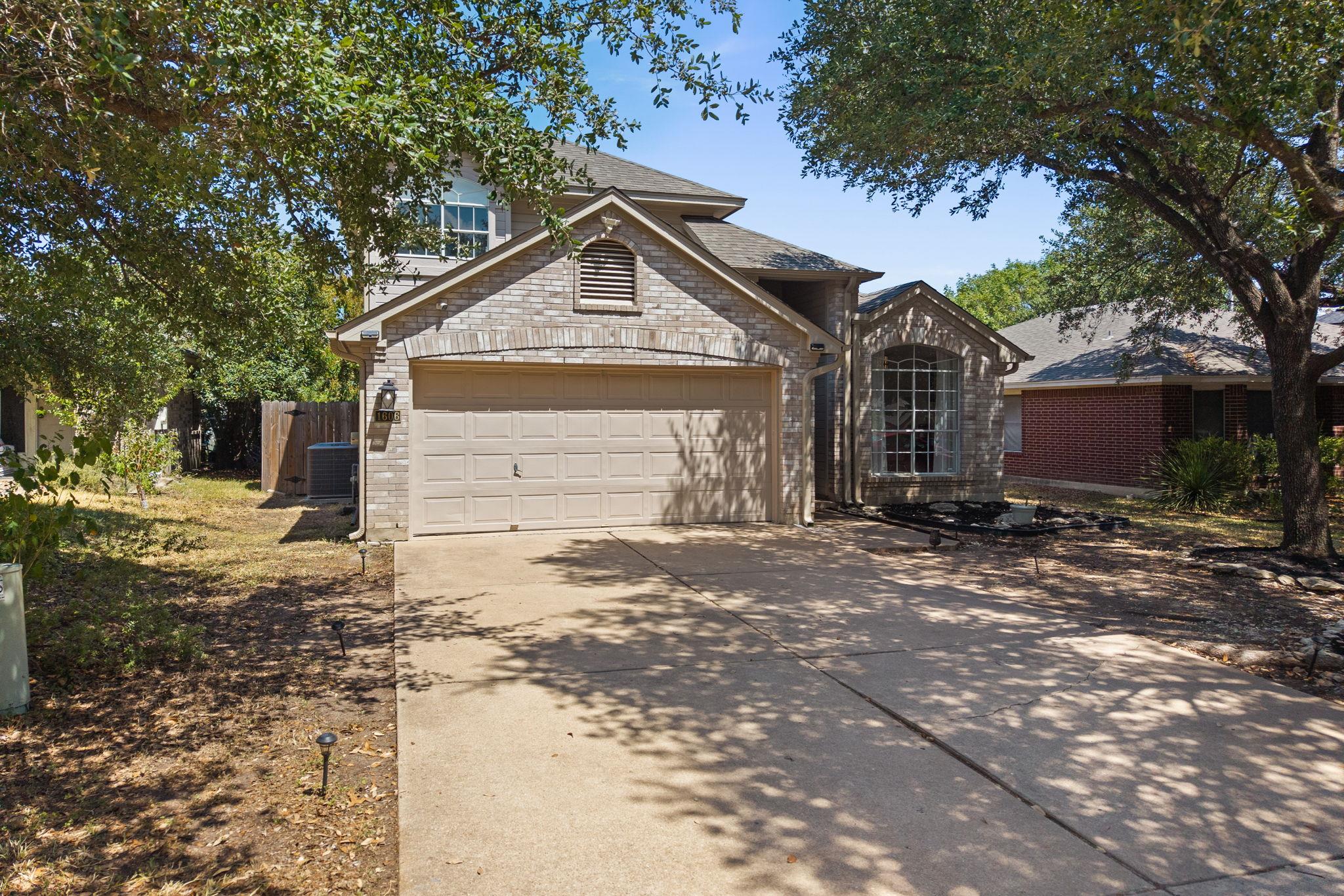 Brick home with tan double garage, arched front window, and tree-shaded driveway.