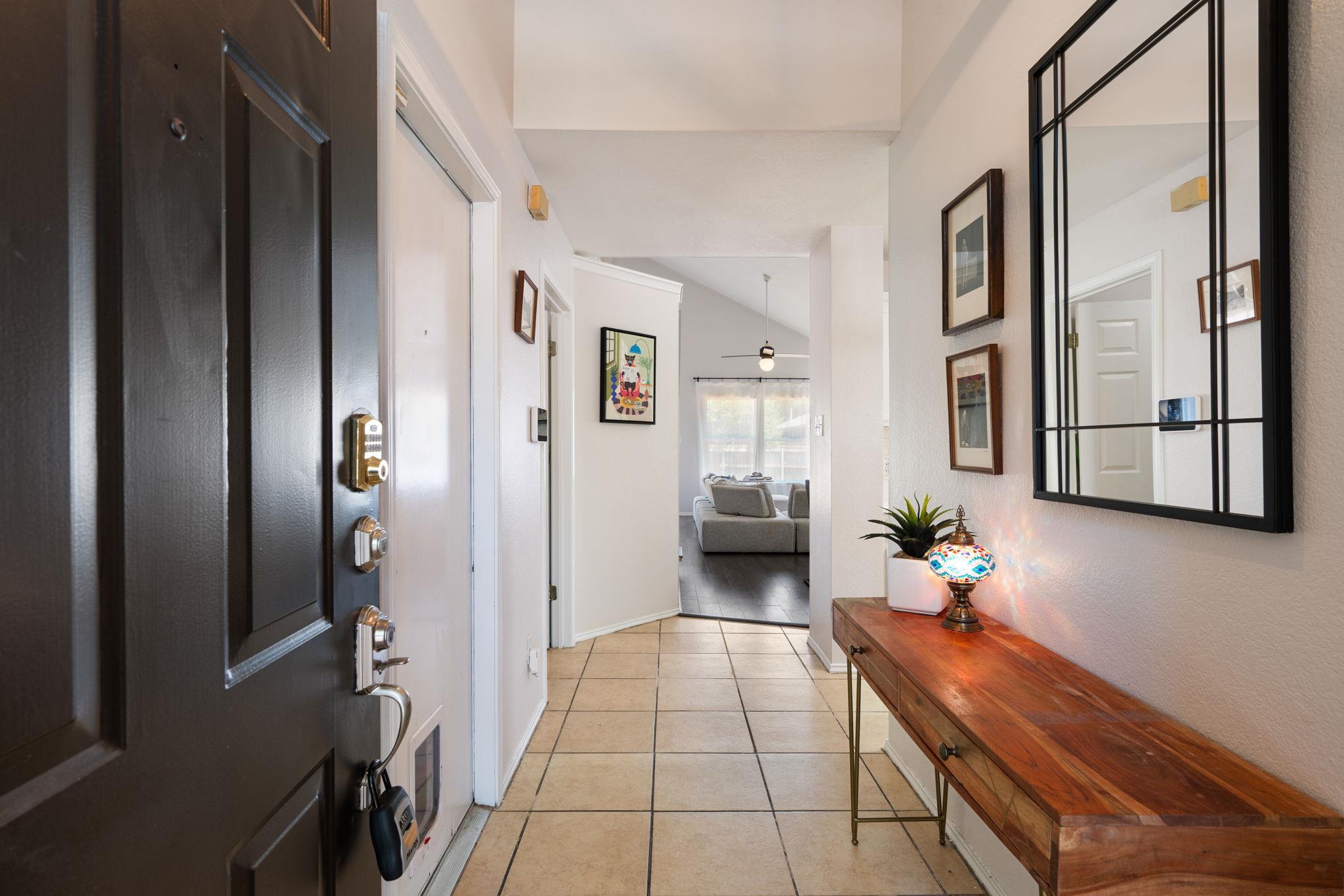 Tiled entryway with a dark front door, wooden console table, colorful lamp, and large mirror.