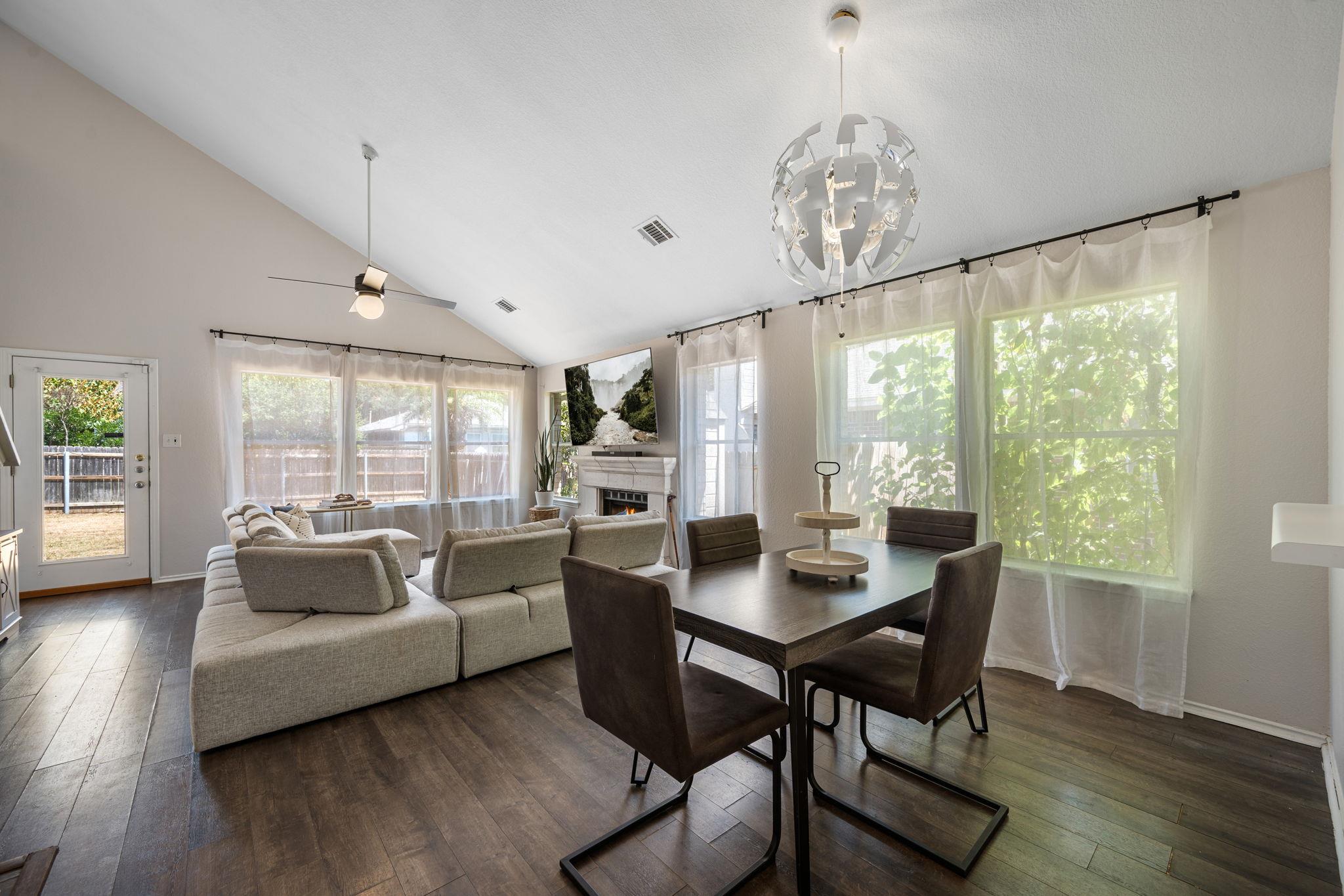 Open-plan living and dining room with vaulted ceiling, large windows, and a fireplace.
