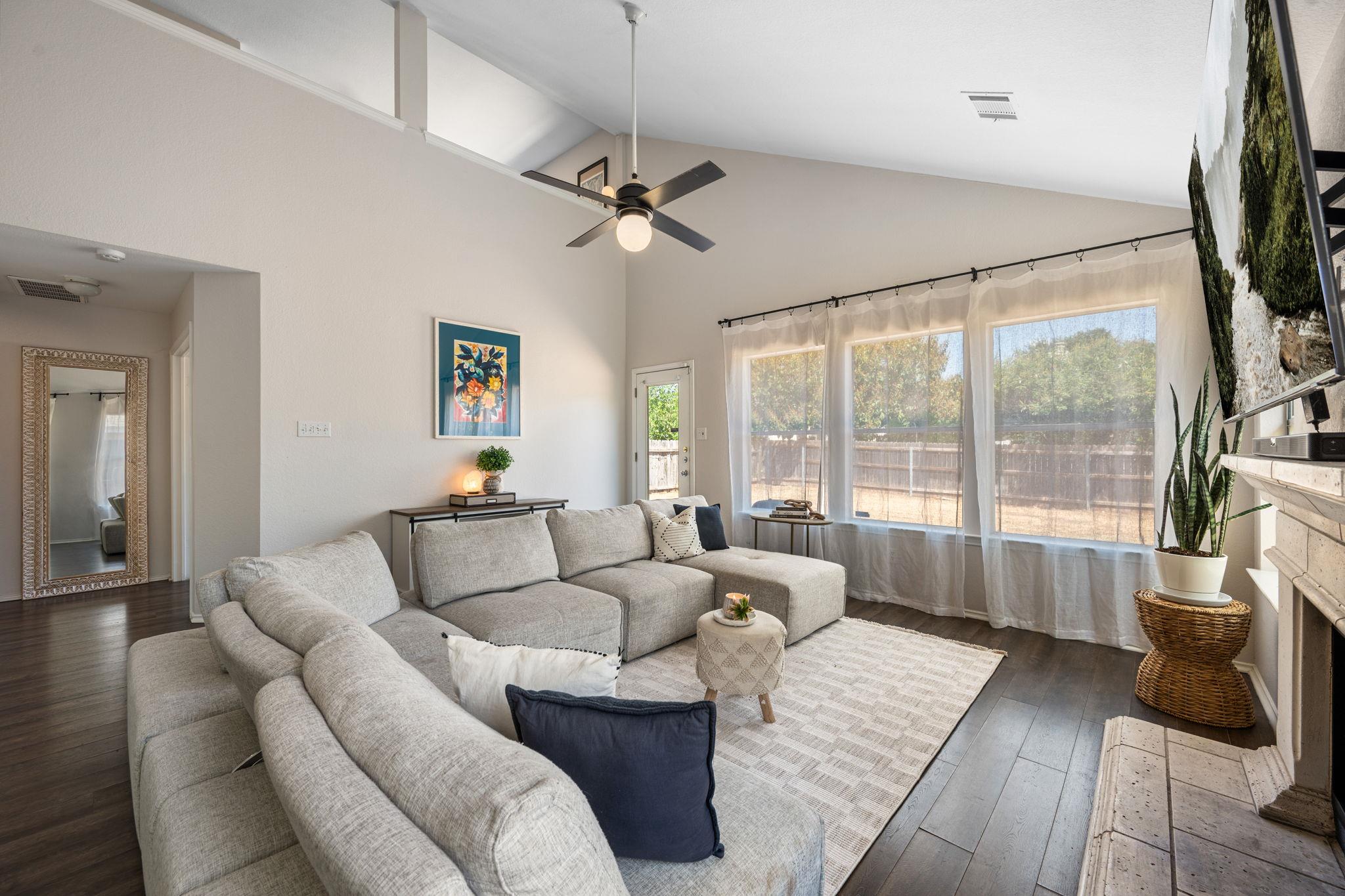 Living room with a gray sectional, vaulted ceiling, and large windows overlooking a fenced backyard.