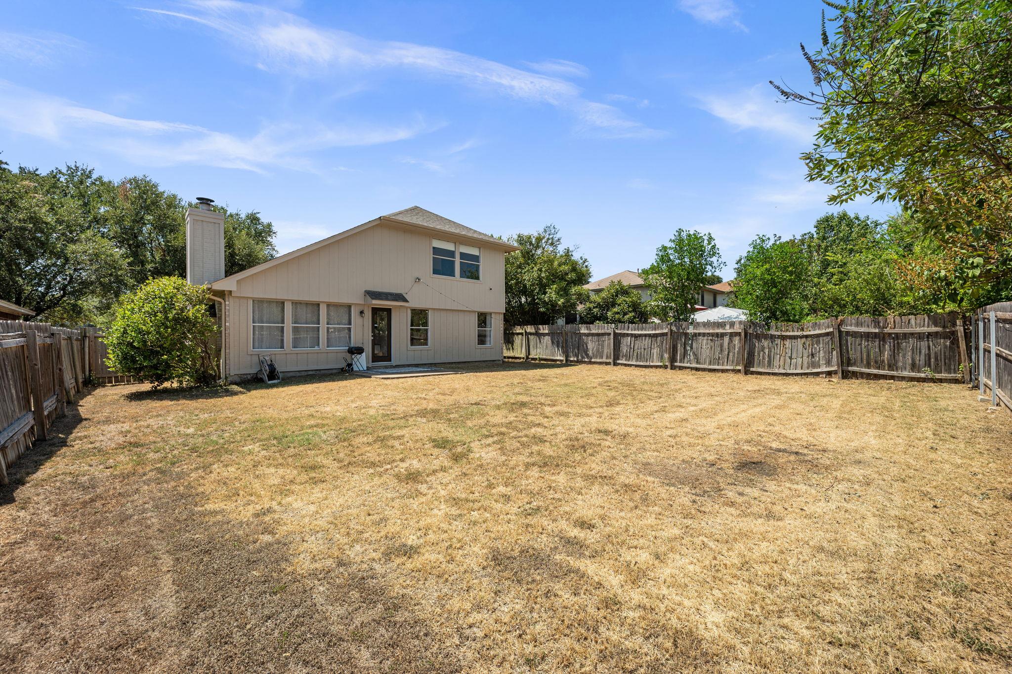 Two-story beige home behind a large fenced backyard with dry grass and mature trees.