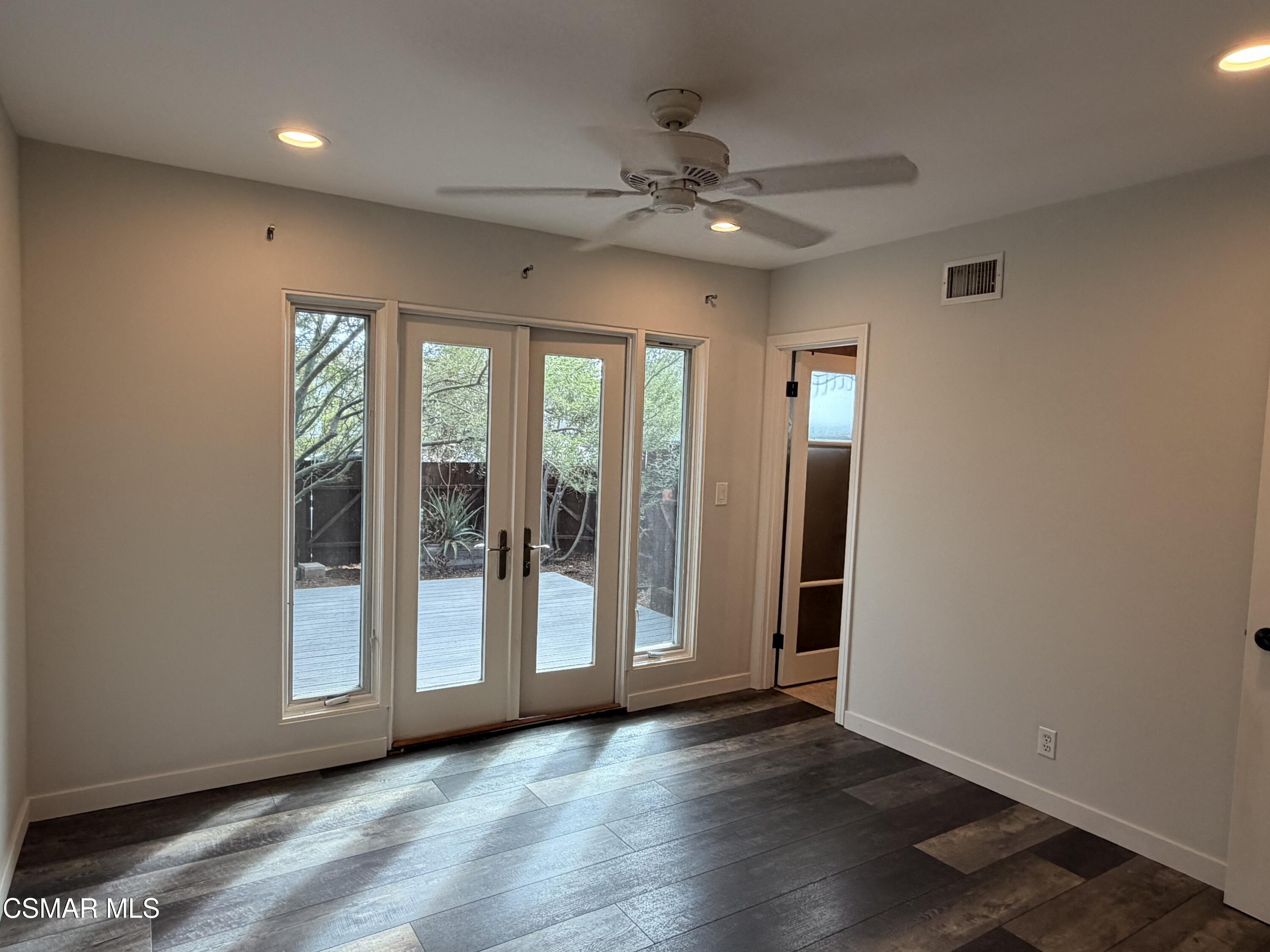 Empty room with dark plank flooring, ceiling fan, and glass doors opening onto a deck.