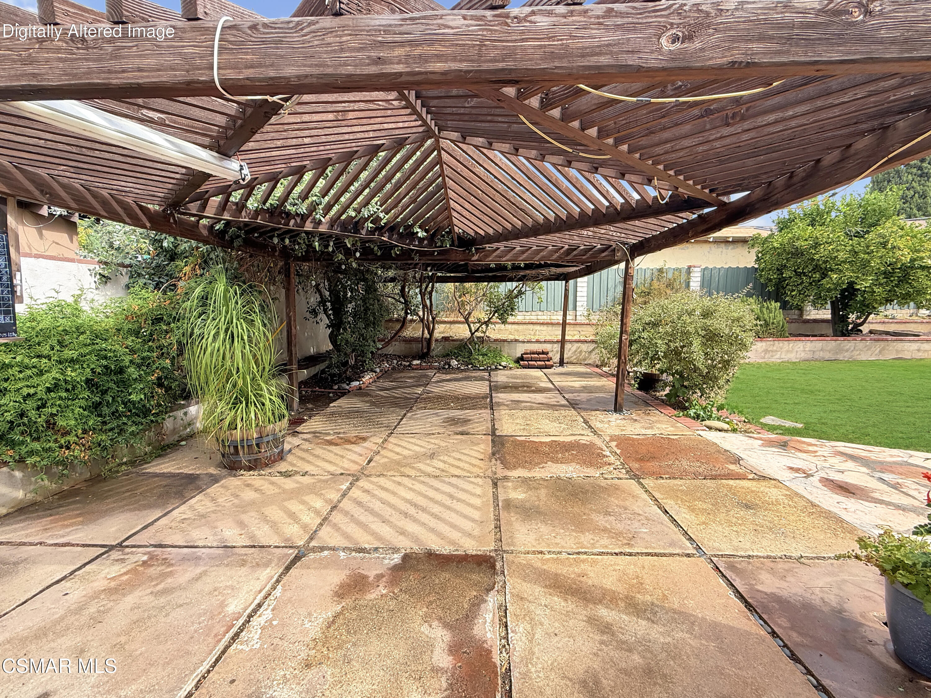 Stone-paved backyard patio beneath a broad wooden pergola, bordered by shrubs and a green lawn.