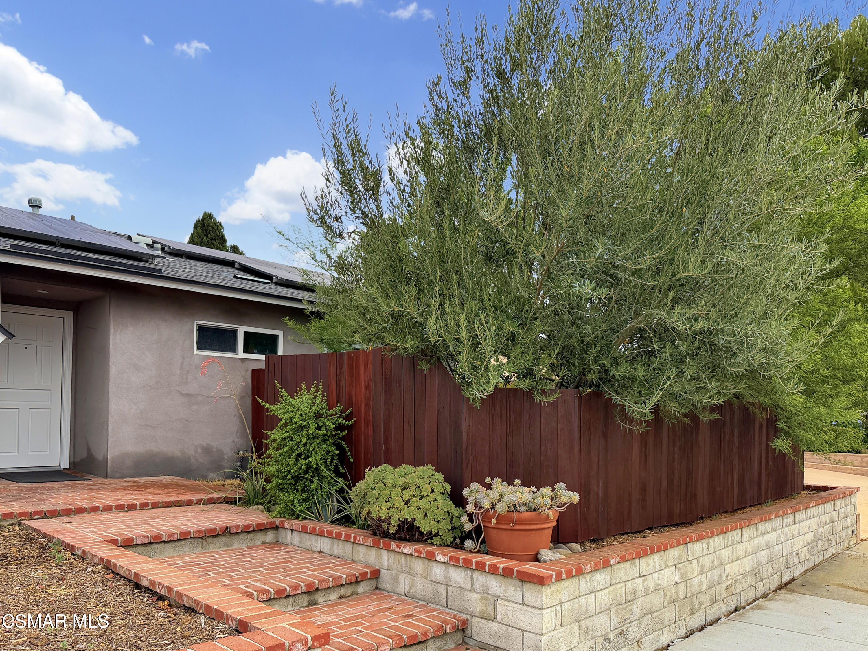 Home entrance with brick steps, raised planter, red fence, and mature tree.