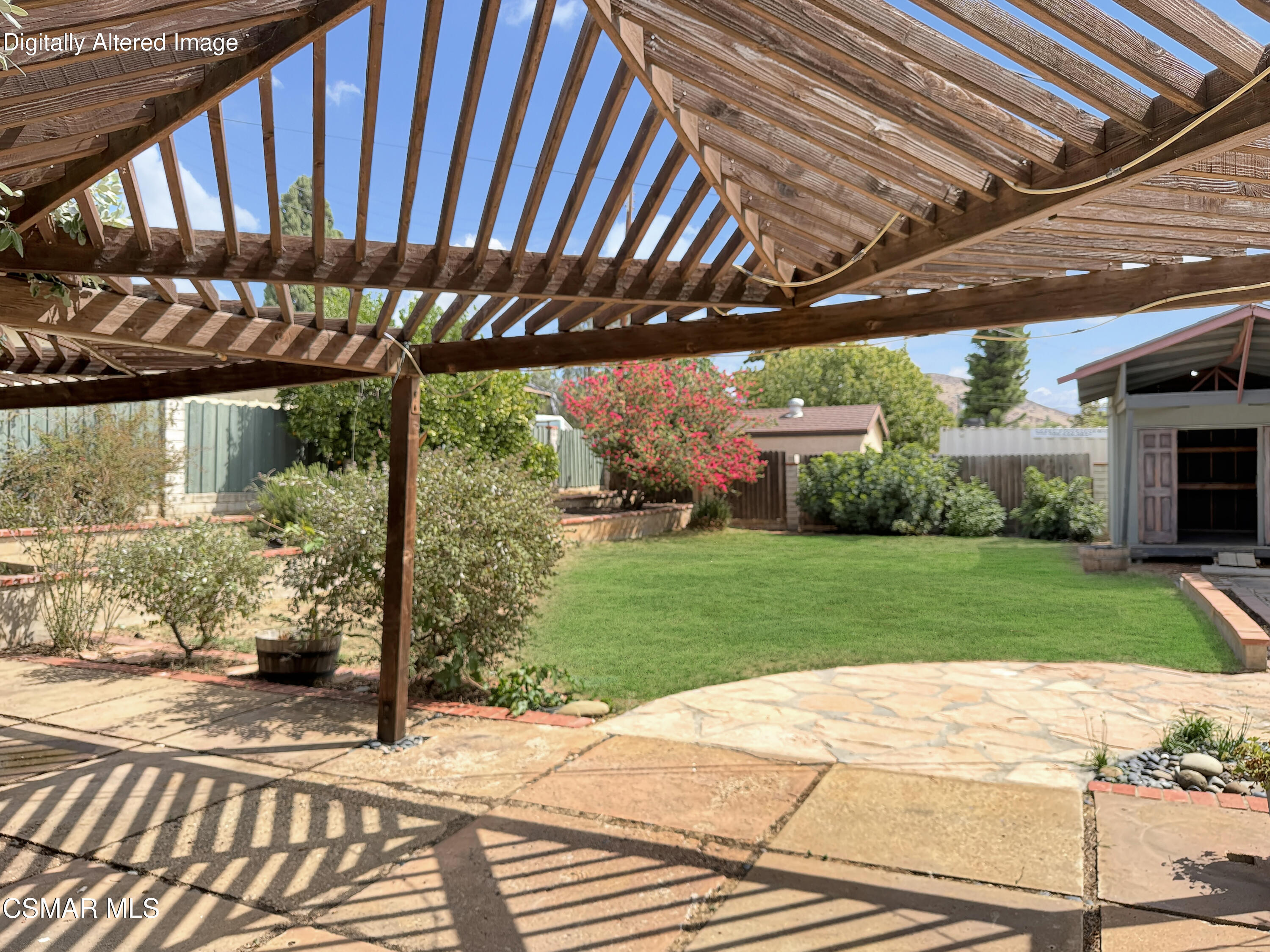 Backyard patio beneath a wooden pergola, with green lawn, flowering shrubs, and garden shed.