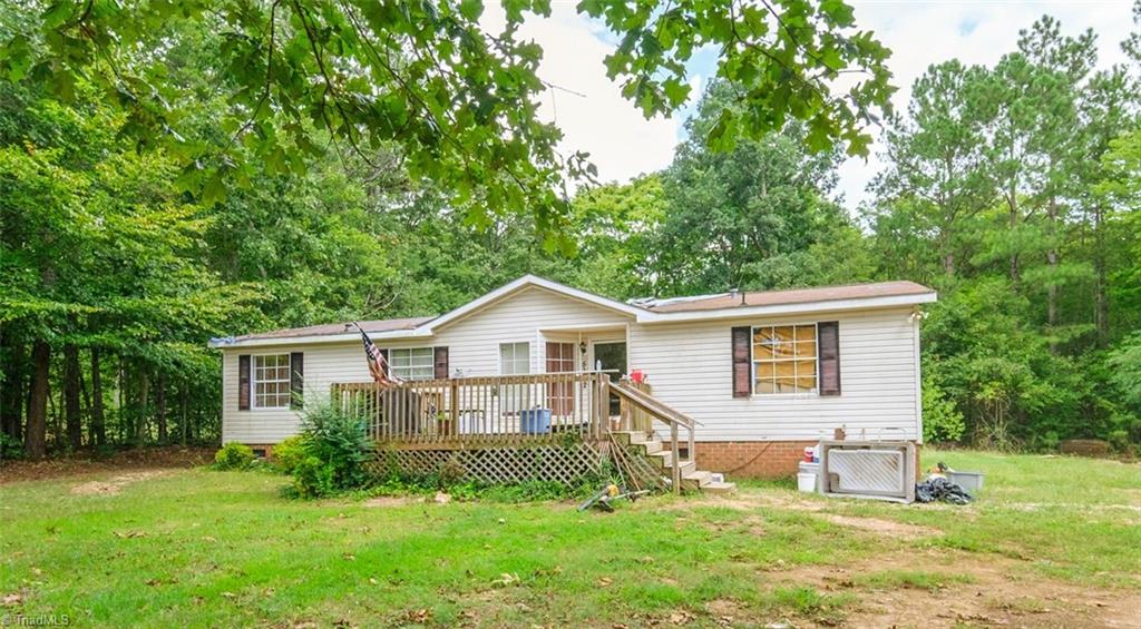 White single-story home with a raised wooden deck in a tree-lined grassy clearing.
