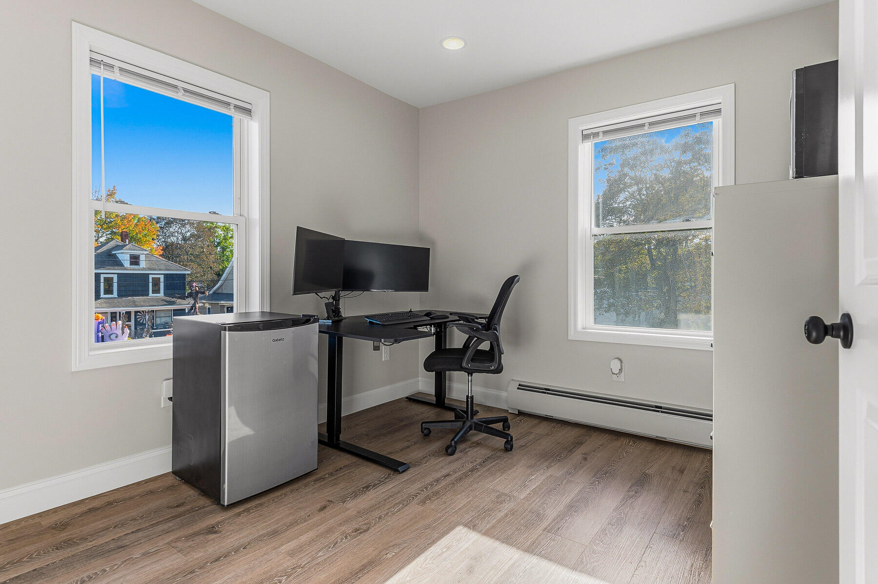 Home office with a black desk, dual monitors, an office chair, and two bright windows.
