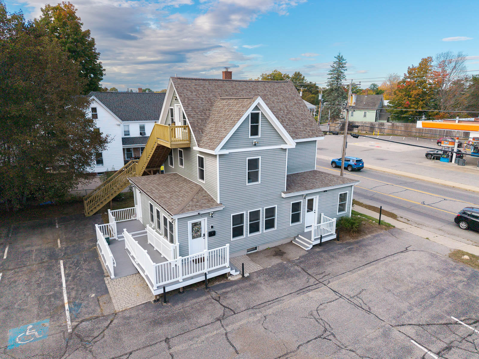 Gray two-story house with white-railed decks and an exterior staircase beside a street and parking lot.