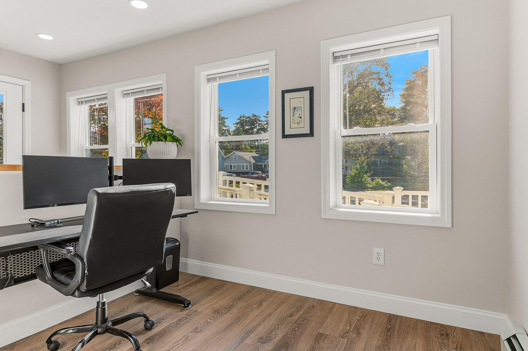 Home office with a dual-monitor desk and windows overlooking autumn trees.