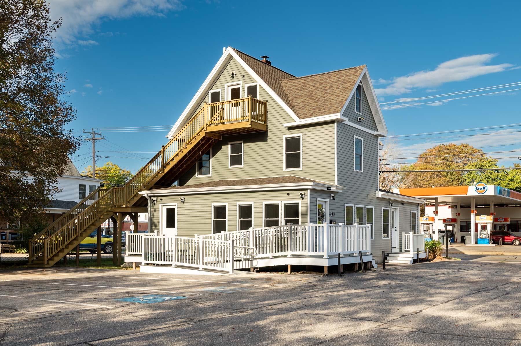 Gray-green building with a long exterior staircase, upper balcony, and white-railed patio beside a Gulf gas station.