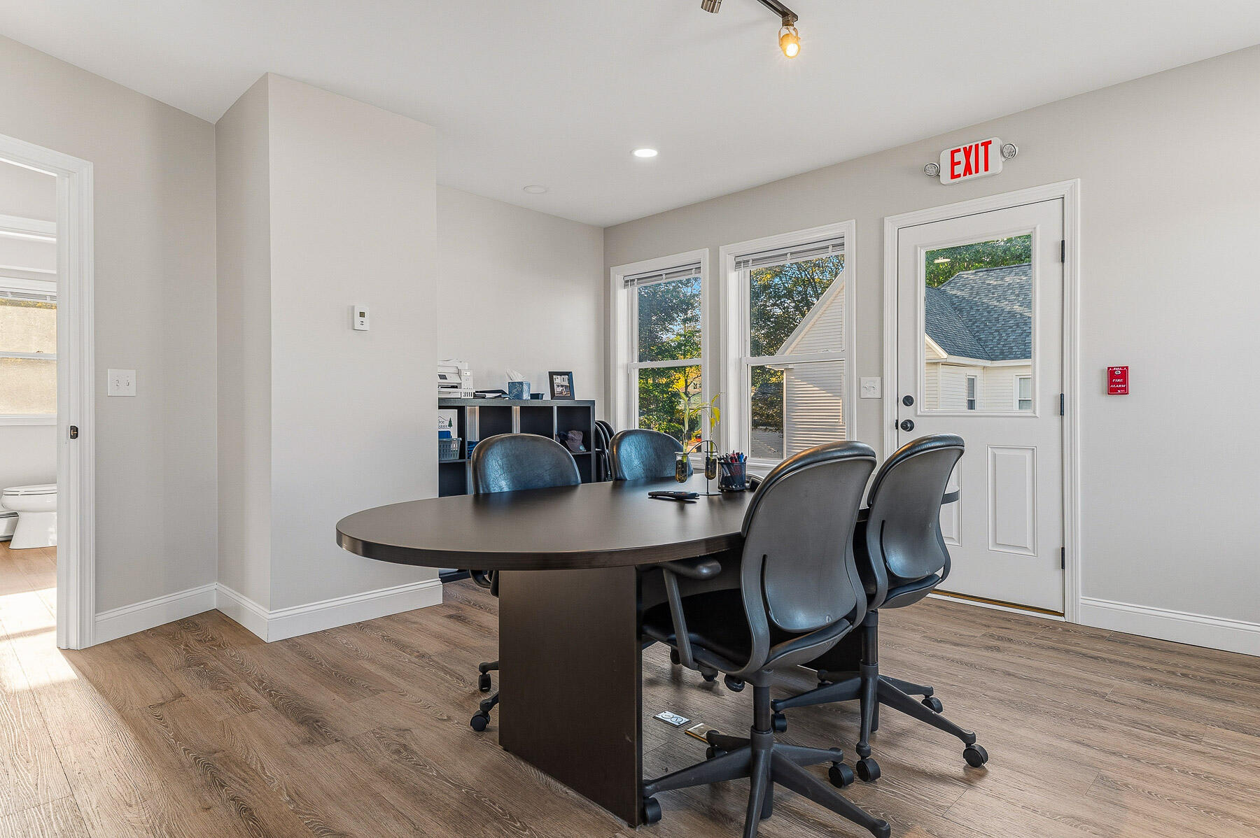 Office meeting room with an oval conference table, rolling chairs, and windows beside an exit door.