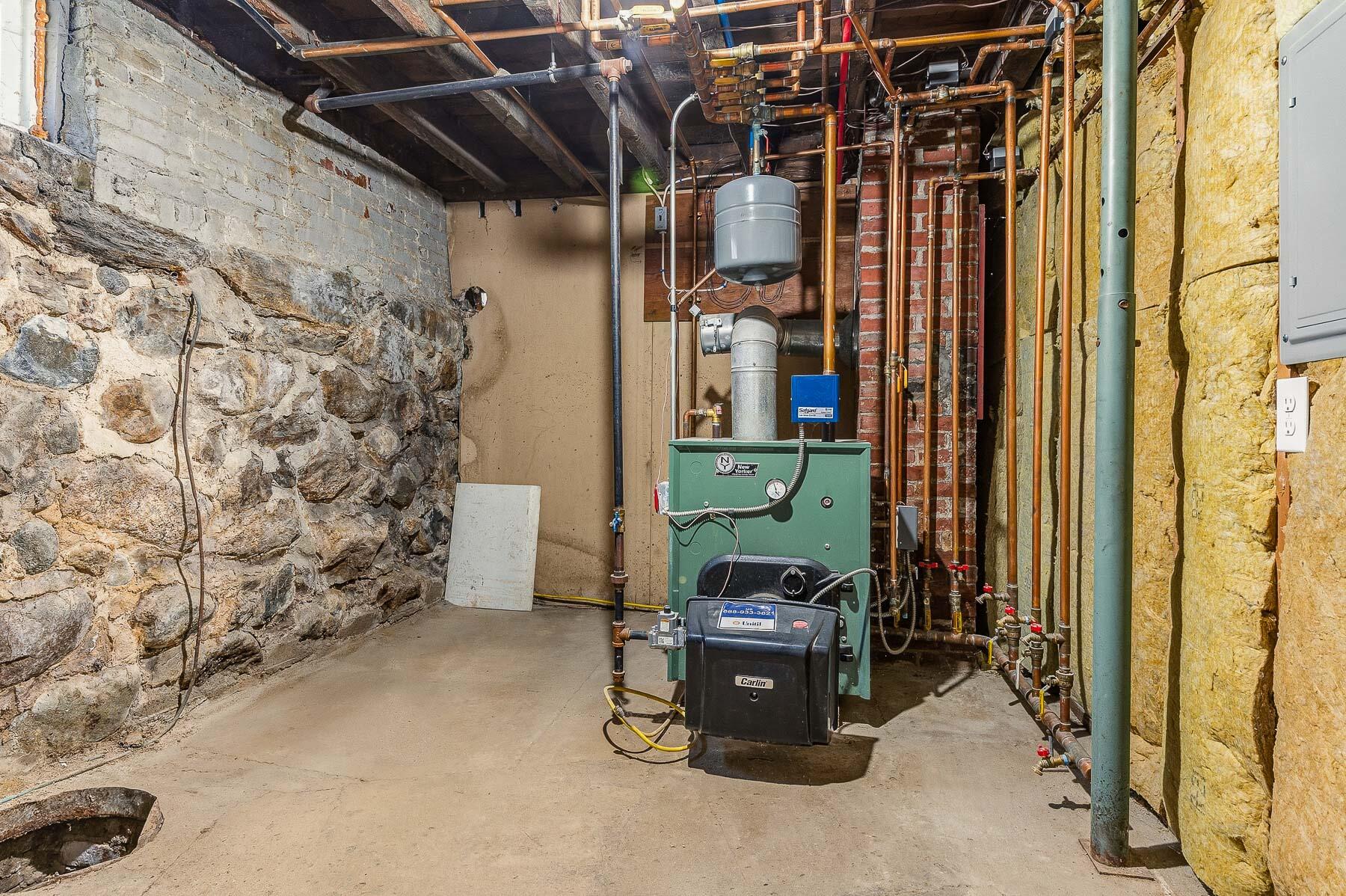 Basement boiler and burner surrounded by exposed pipes beside a rough stone foundation wall.