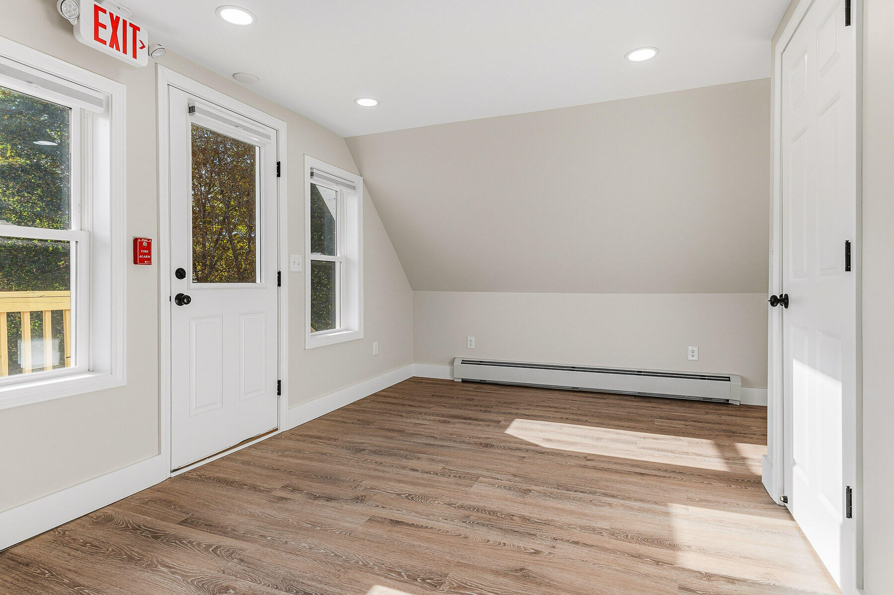 Vacant interior room with wood-look floors, recessed lights, and windows beside a glazed exterior door.