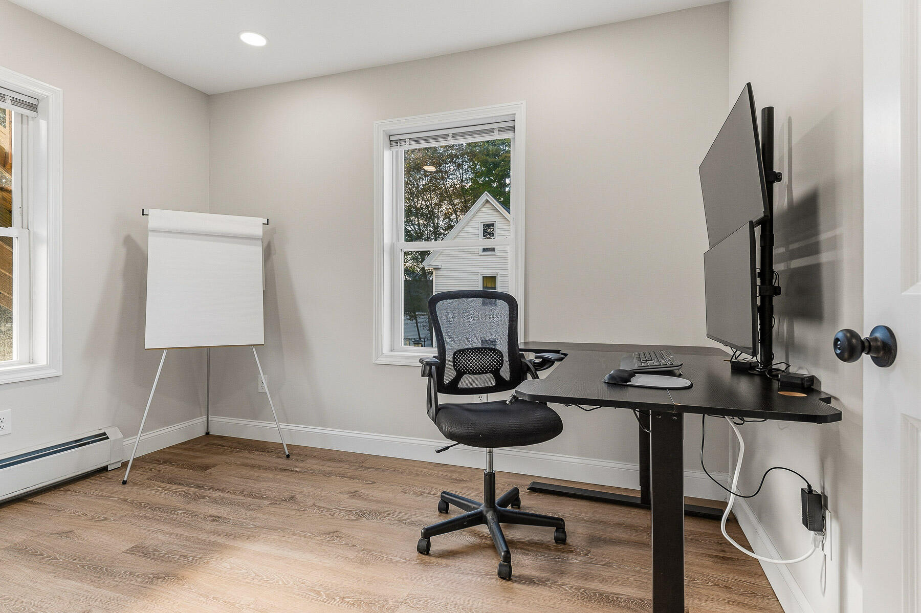Home office with a black desk and monitors, mesh chair, blank flip chart, and two windows.