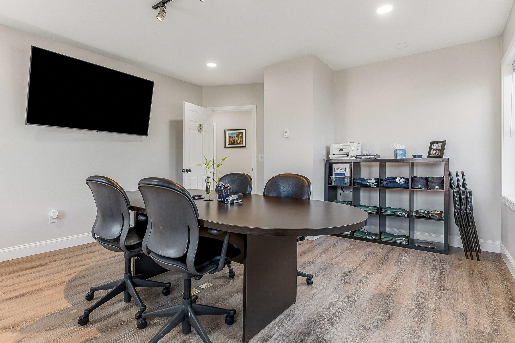 Office conference room with a large table, rolling chairs, wall-mounted television, and shelving.