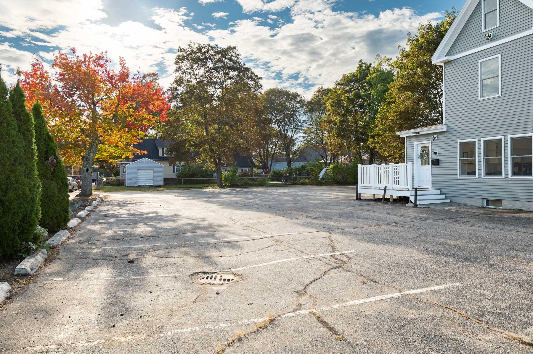 Gray-sided building with a white entrance deck beside a large paved parking lot and autumn trees.