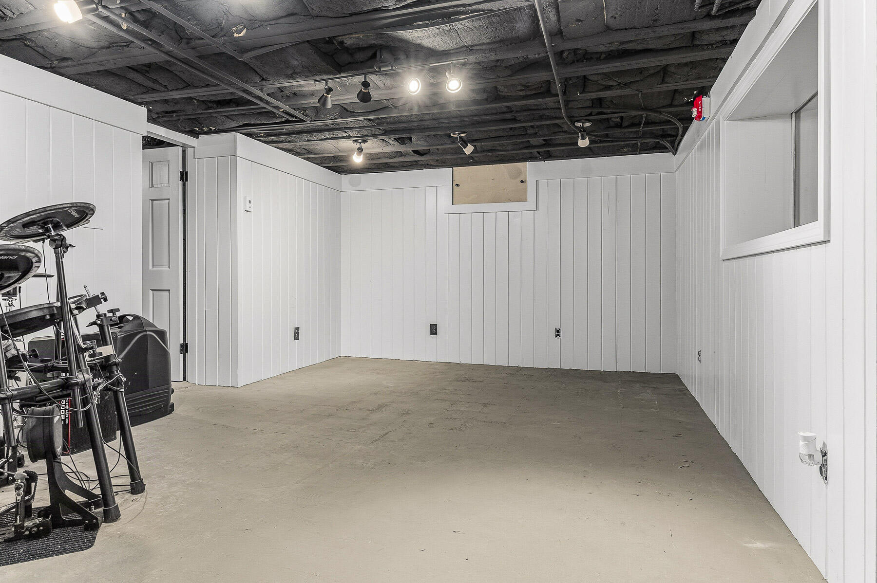 Basement room with white paneled walls, concrete floor, exposed ceiling, and exercise equipment along one side.
