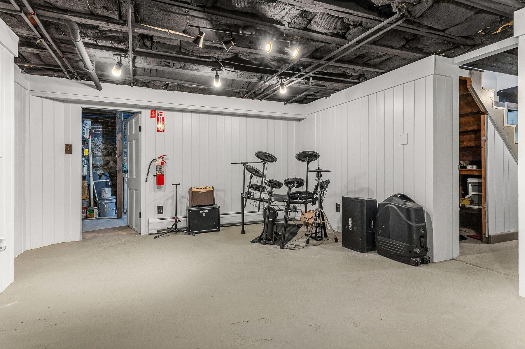 Basement music room with an electronic drum kit, speakers, exposed ceiling, and stairs.