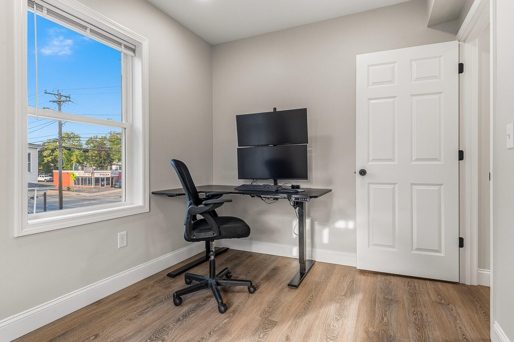 Home office with a dual-monitor desk and chair beside a large street-facing window.