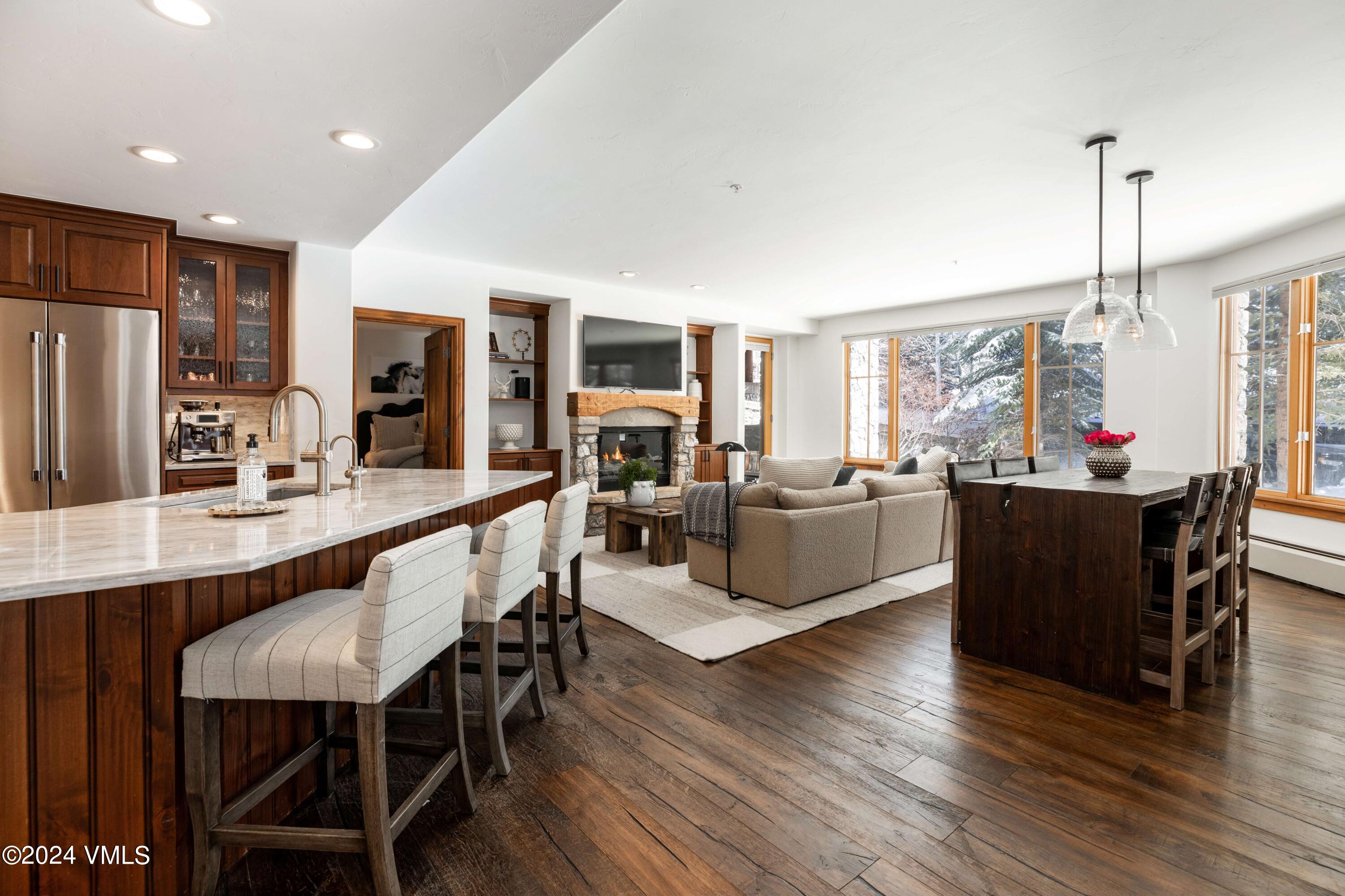 Open-plan kitchen and living room with marble island, stone fireplace, dining table, and large windows.