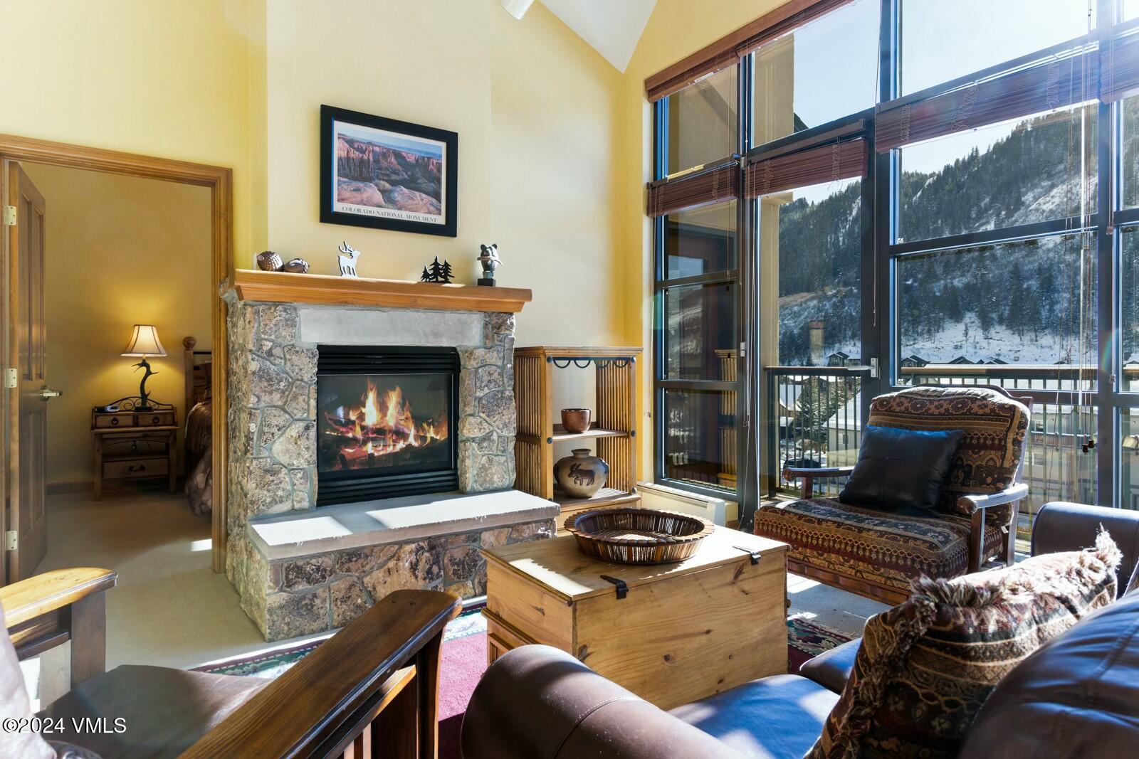Living room with a stone fireplace and tall windows overlooking a snowy mountain.