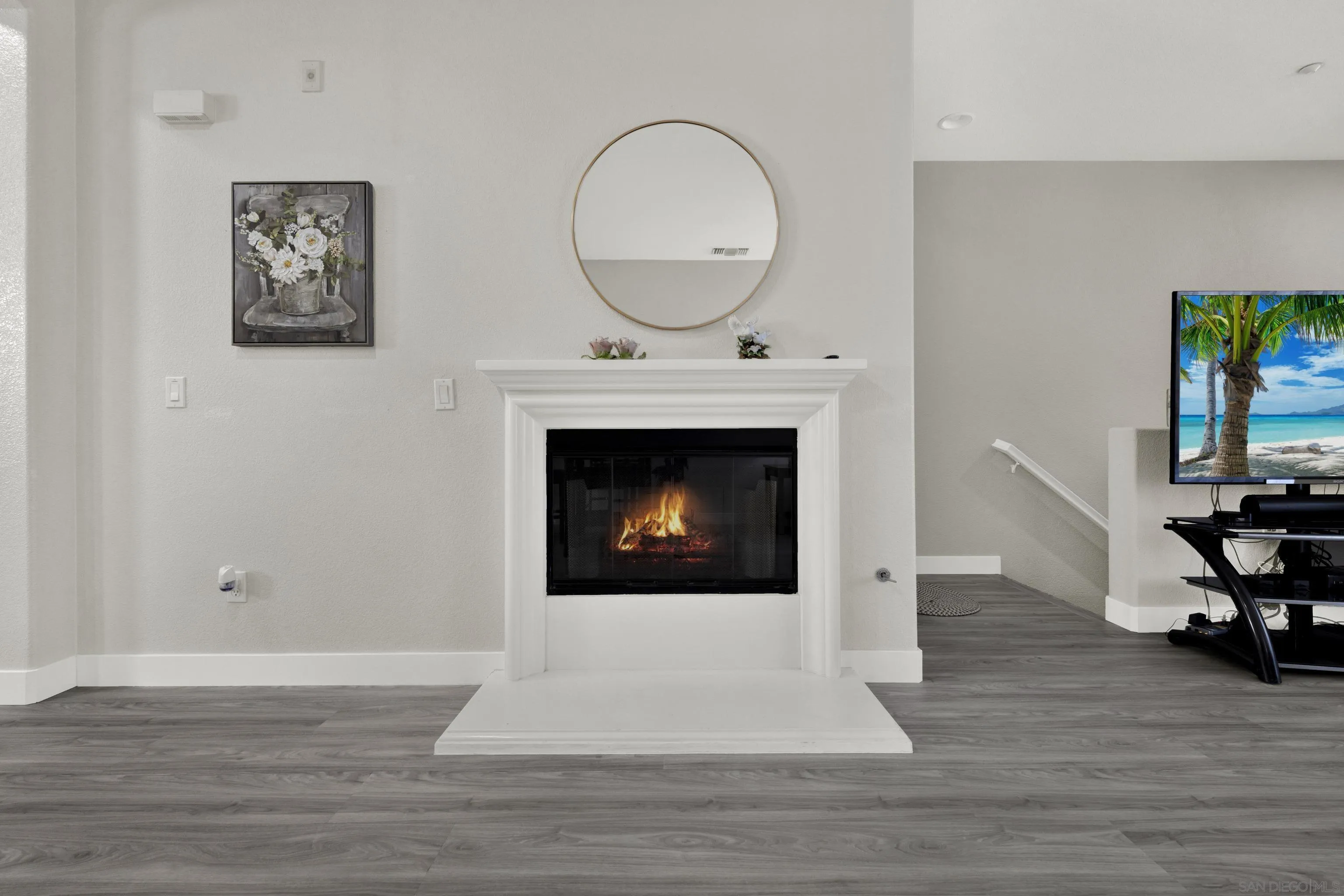 Living room with a lit white fireplace, round mirror, and gray wood flooring.