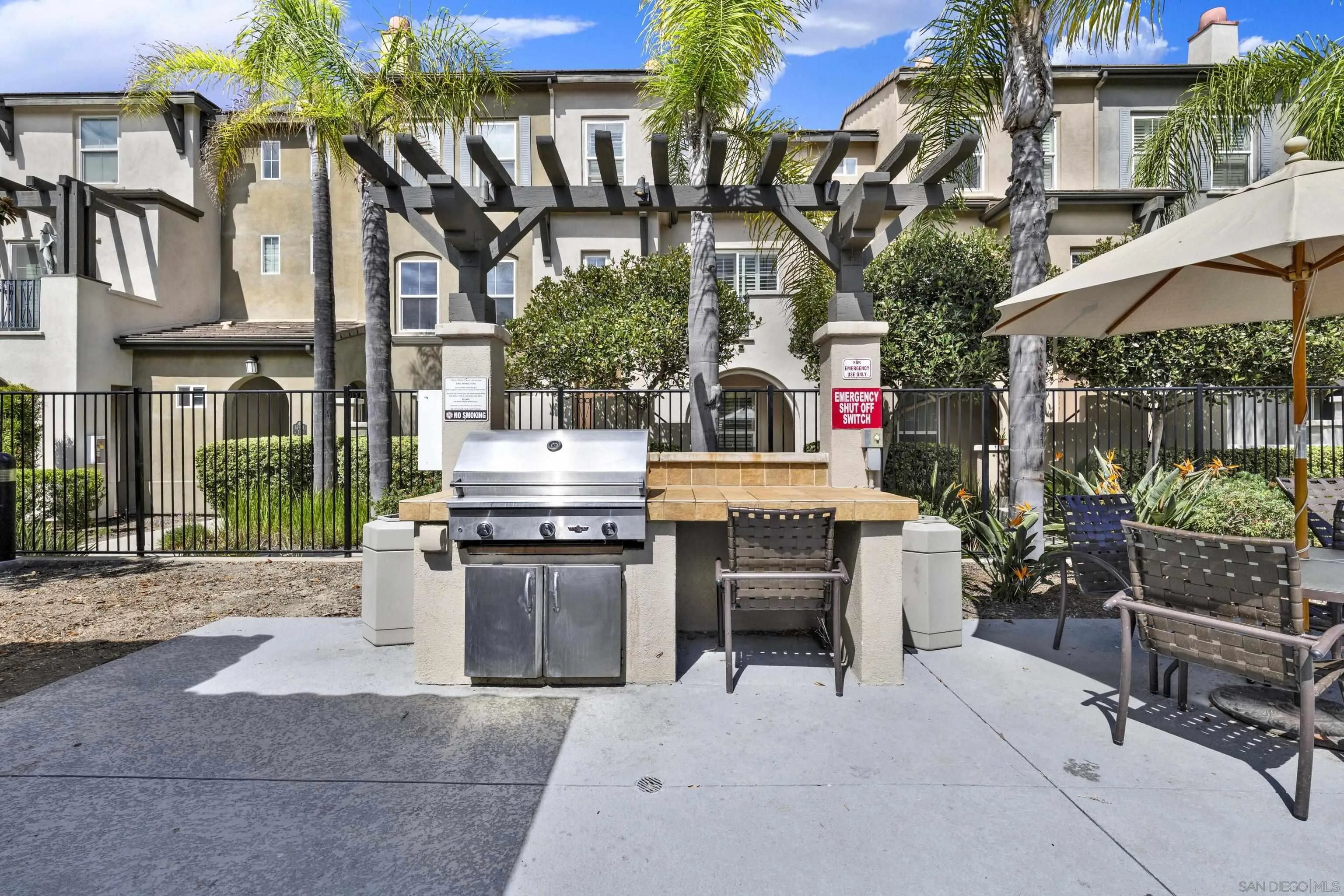 Outdoor barbecue area with a stainless-steel grill, pergola, patio seating, and palm trees.