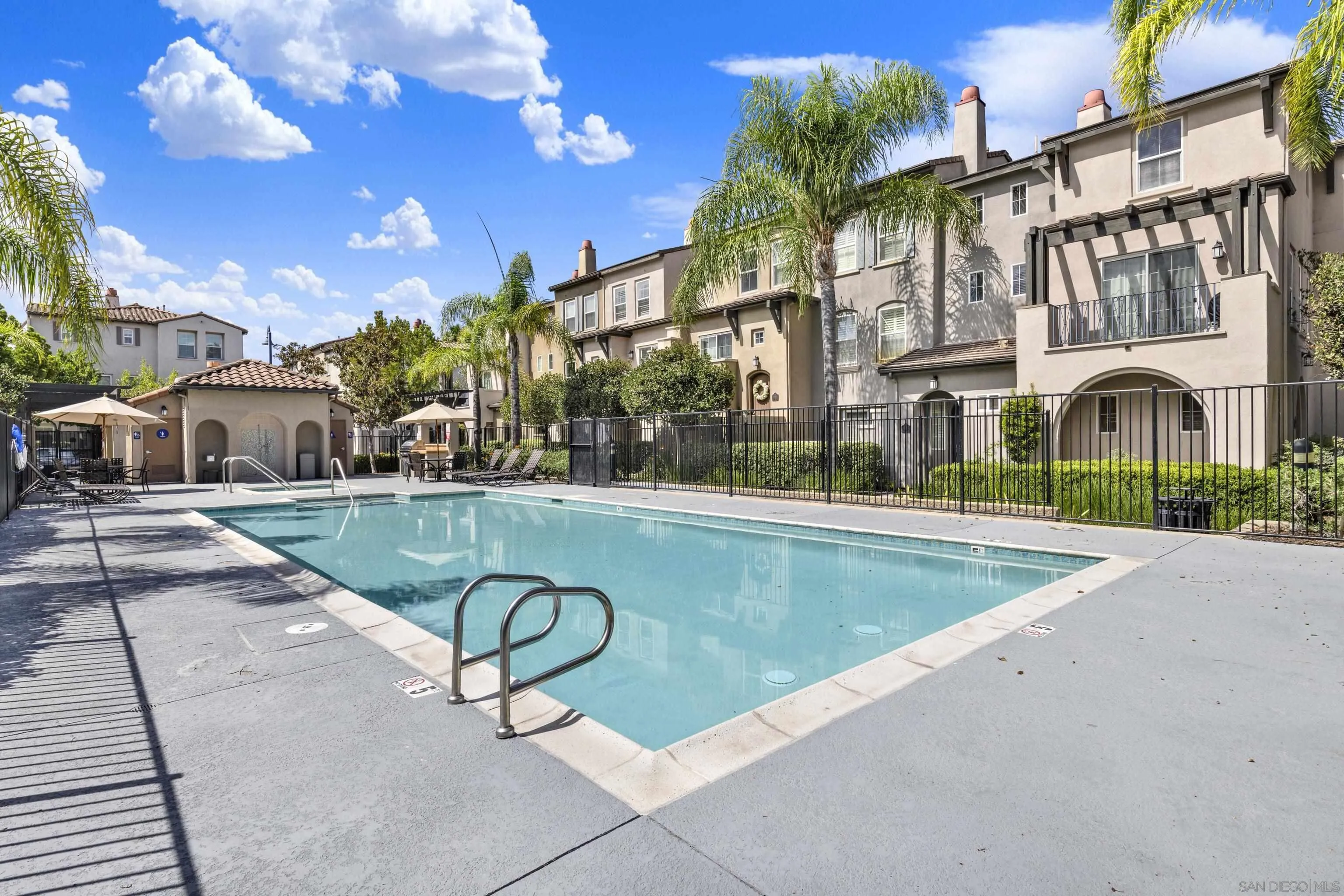 Community pool with a metal ladder, patio umbrellas, and multi-story homes in the background.